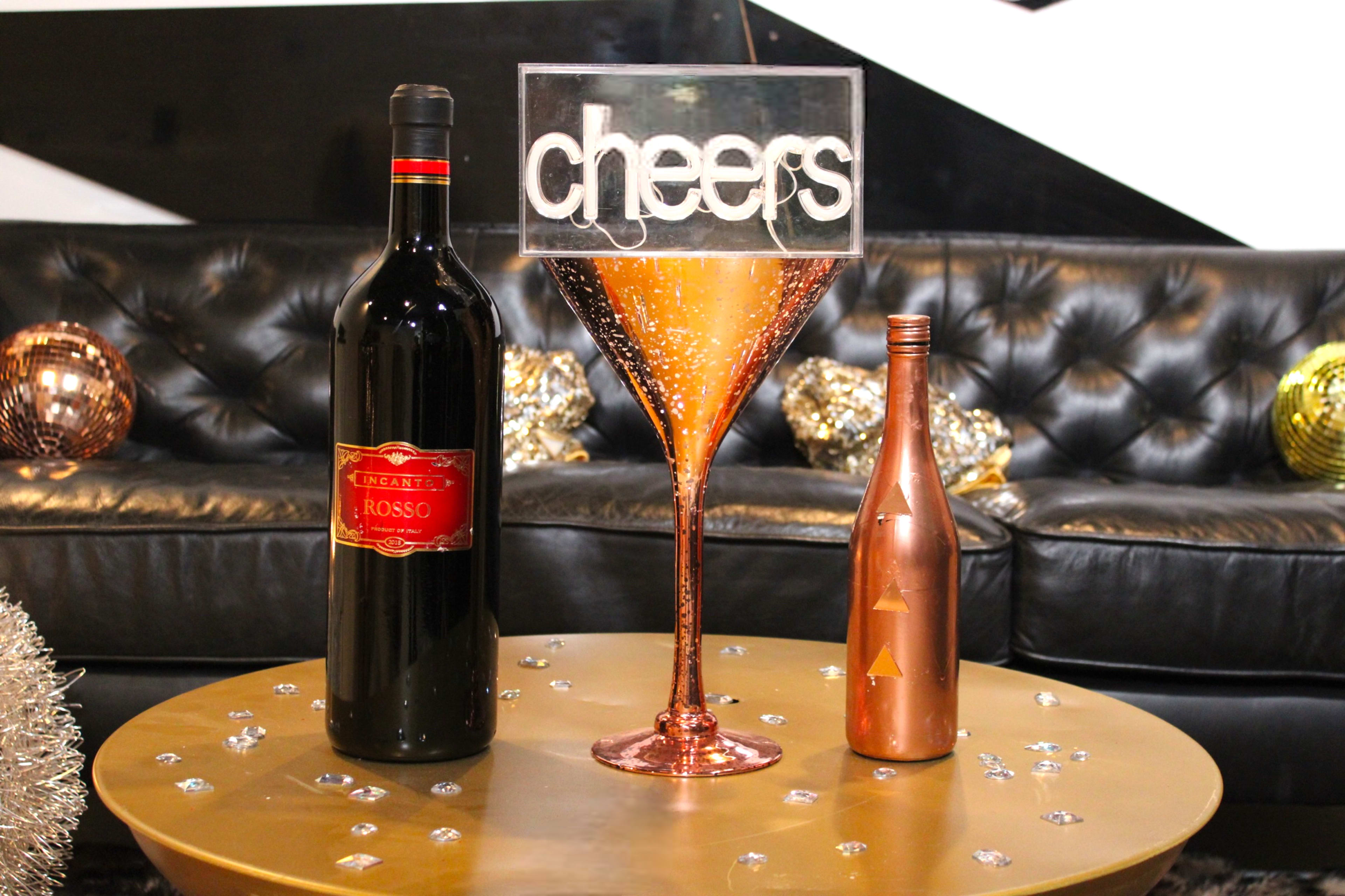 A large decorative martini glass filled with glimmering liquid stands on a gold table, surrounded by a bottle of red wine and a smaller bottle, against a backdrop of a black leather couch and metallic decor.