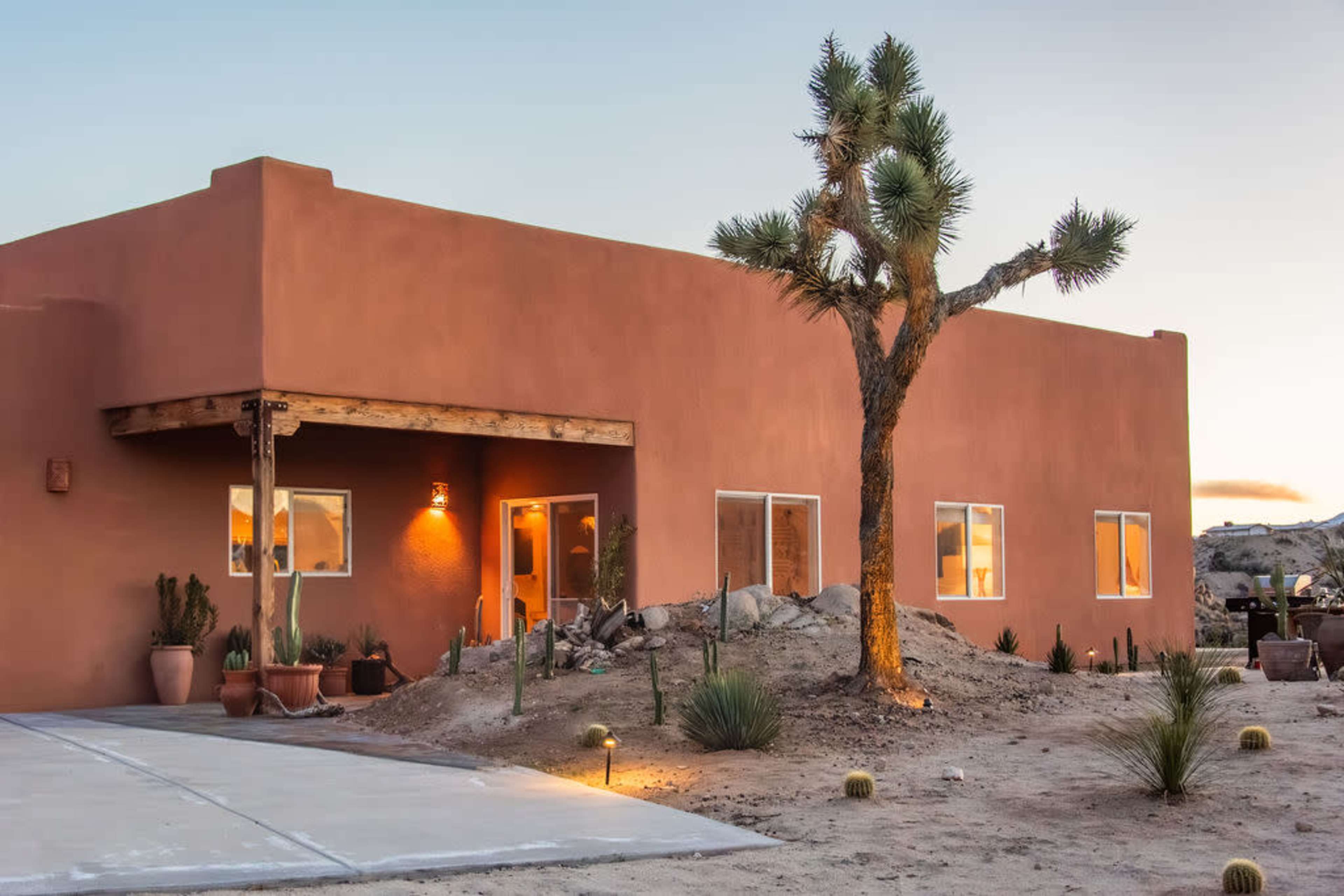 A single-story adobe-style house with large front windows is situated in a desert landscape, featuring a Joshua tree and various potted plants around the exterior.