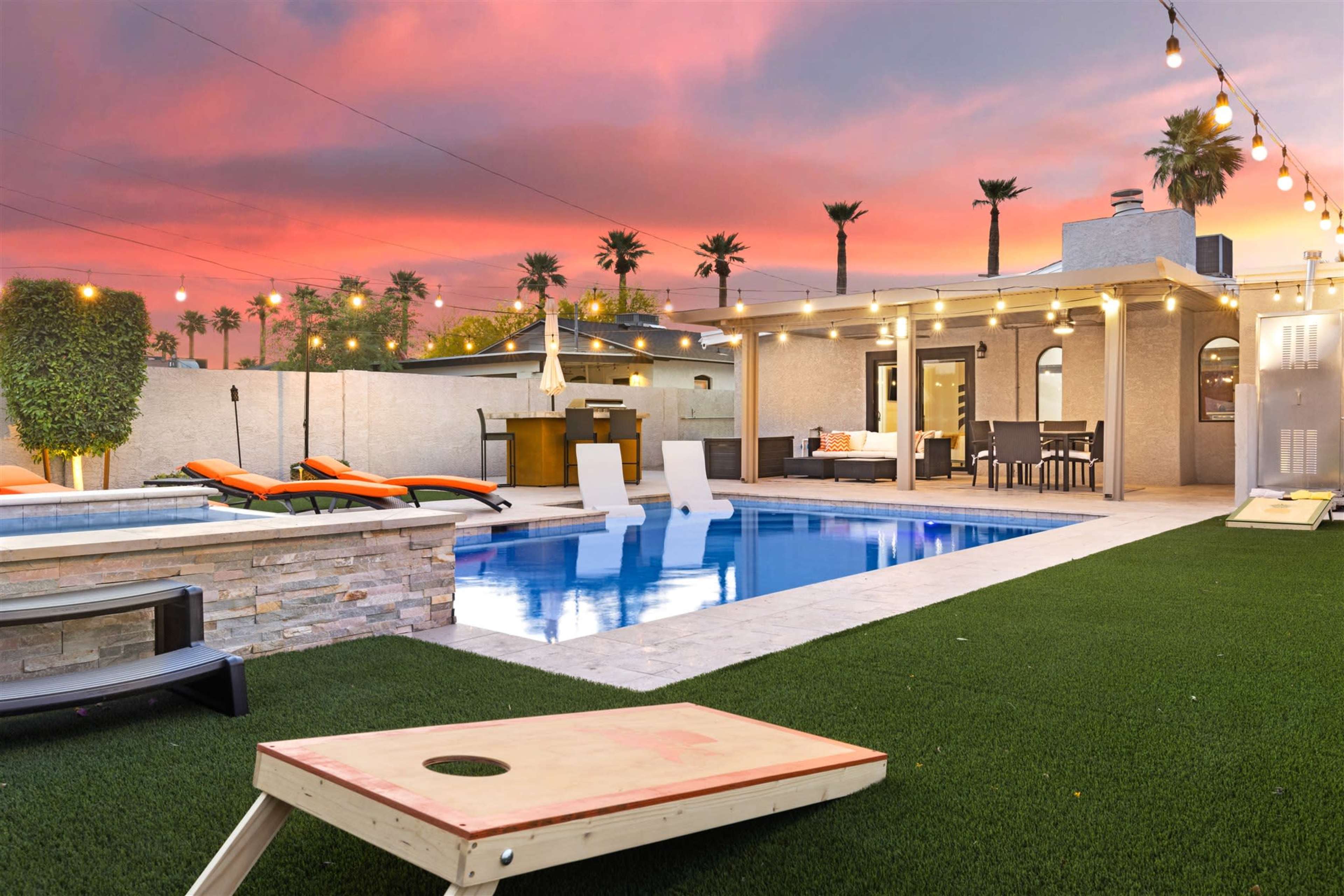 A backyard pool area with lounge chairs, a green lawn, and string lights against a sunset sky.