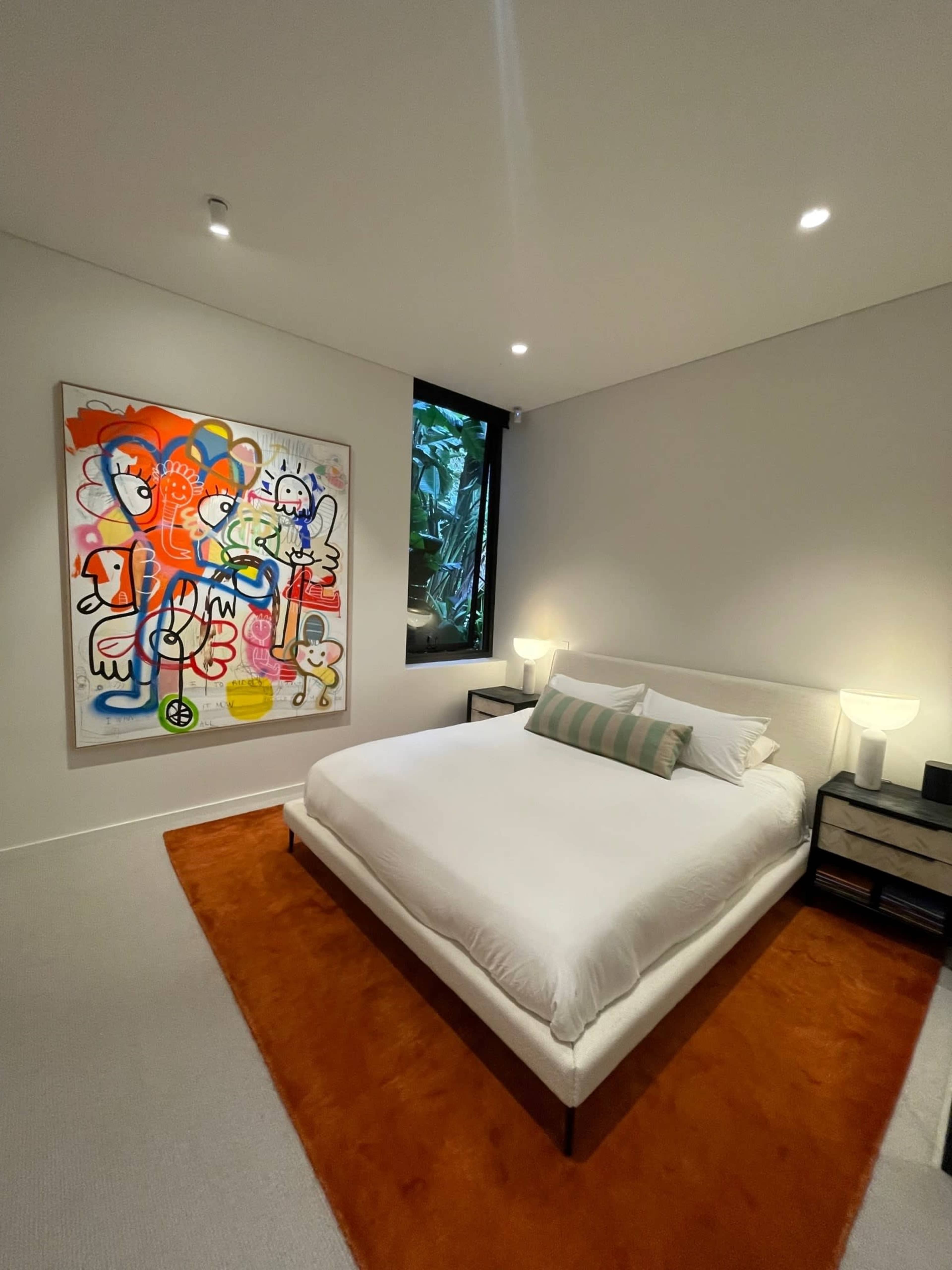 A modern bedroom features a white bed with a geometric orange rug and a colorful abstract painting on the wall.