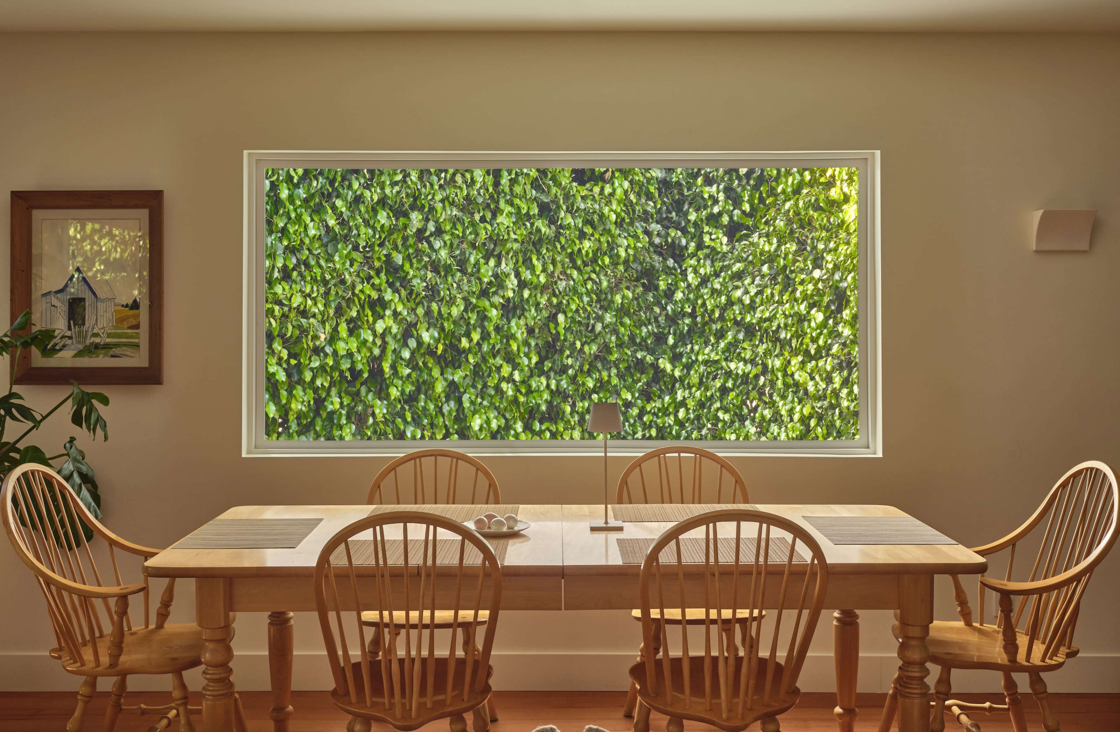 A dining room features a wooden table surrounded by six chairs, with a large window showcasing a green hedge outside.