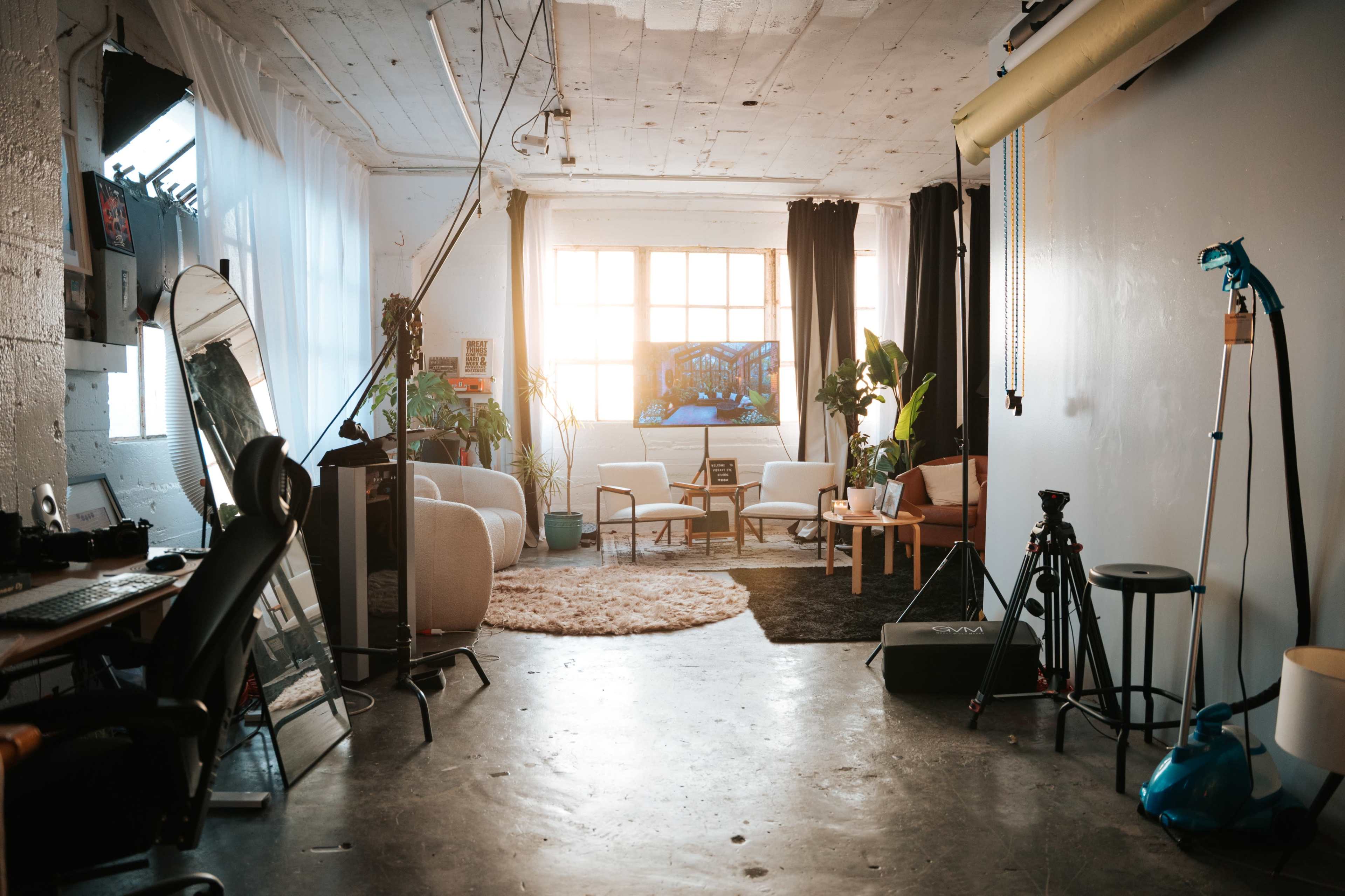The image shows a spacious, well-lit studio with a mix of furniture, plants, and photography equipment arranged throughout the room.