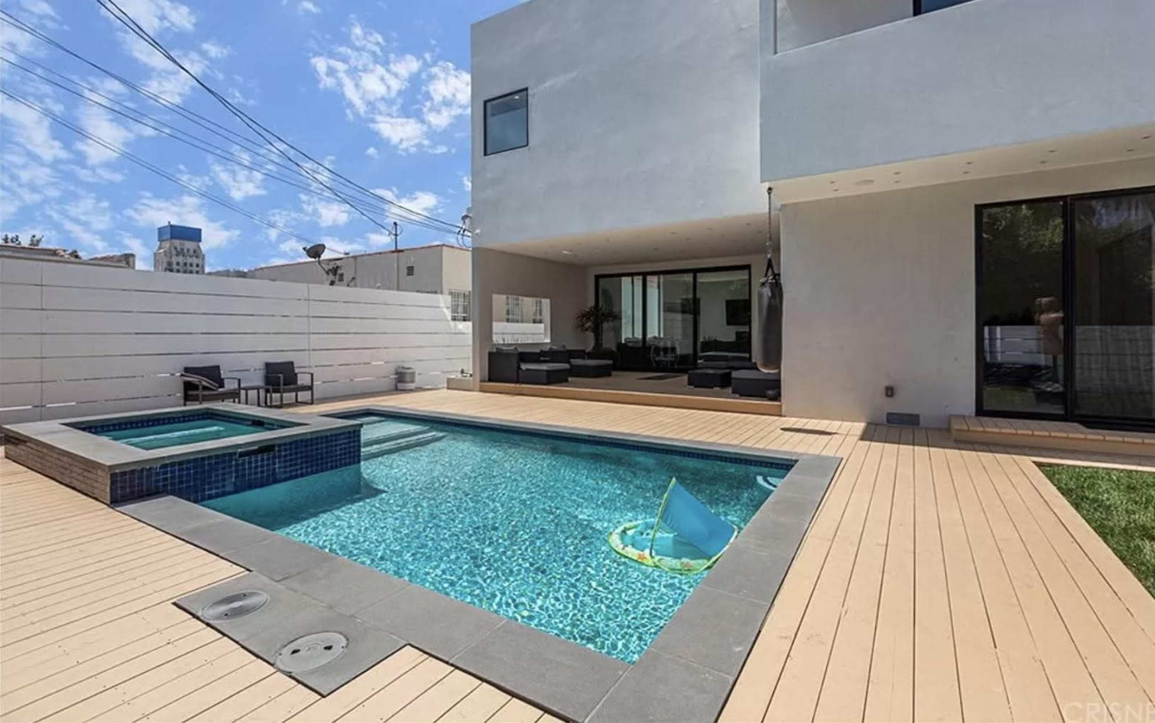 A rectangular swimming pool with a spa is surrounded by wooden decking and features a small inflatable float in the water, with a modern house visible in the background.
