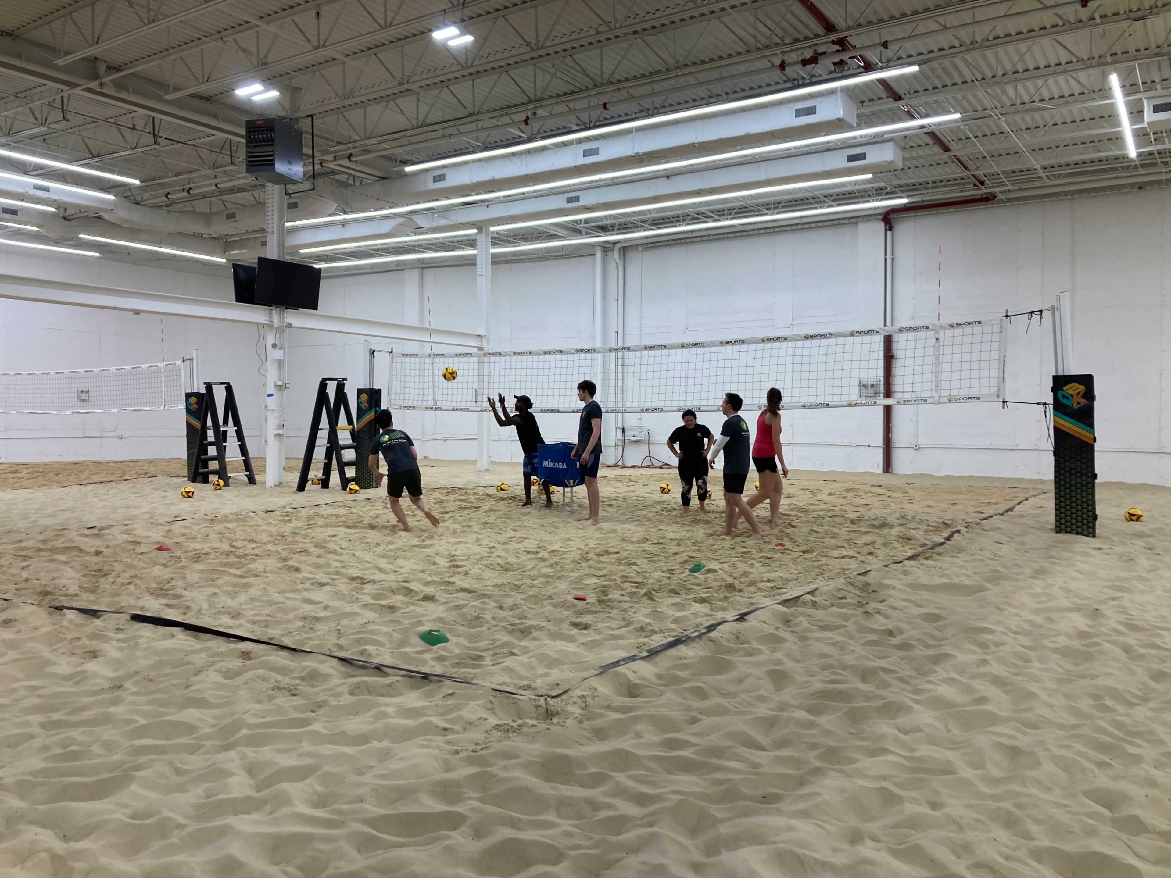 A group of individuals is playing volleyball on a sand court inside a facility with a high ceiling.