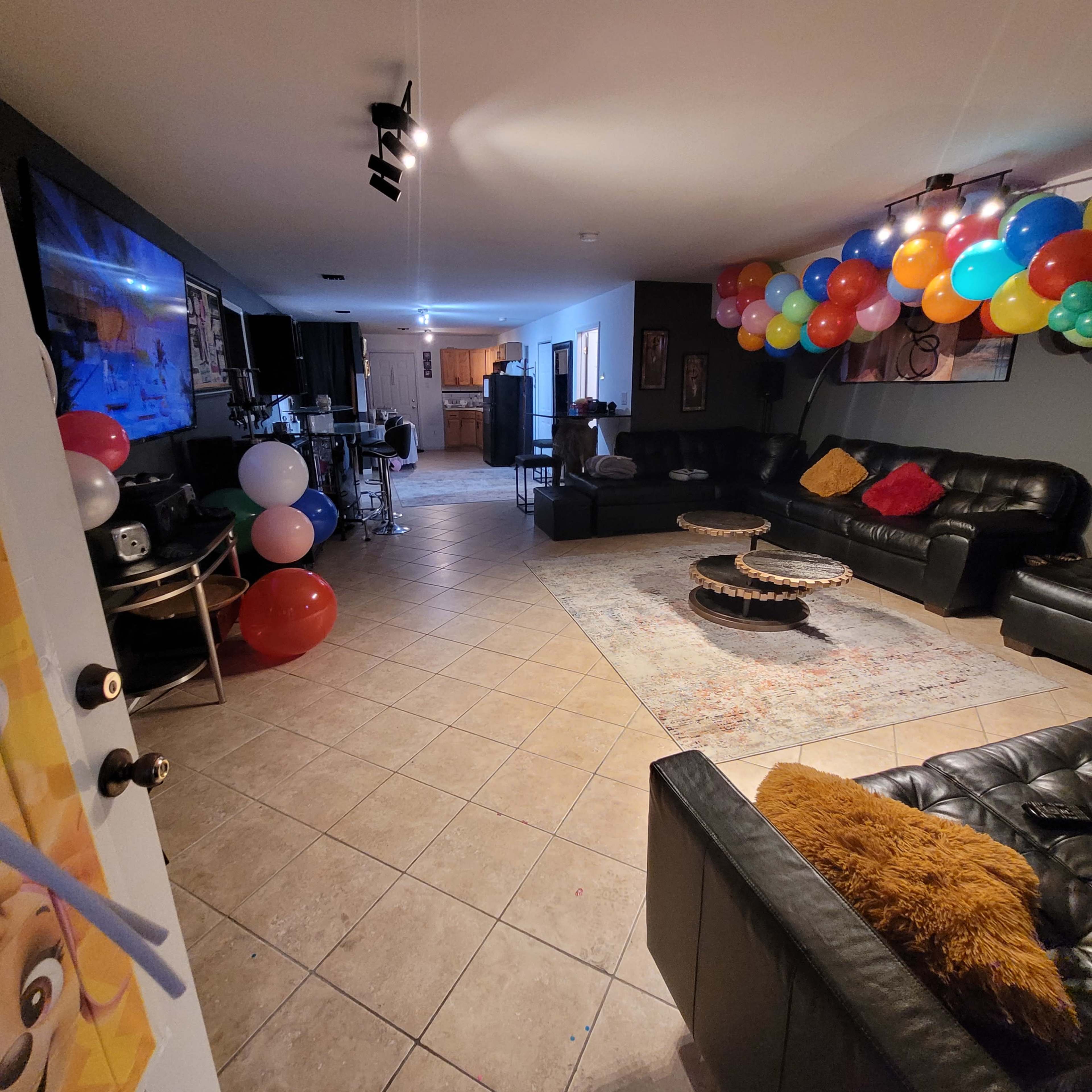 The image shows a spacious basement living area with black leather sofas, a large TV, colorful balloons along one wall, and a rug on the tiled floor.