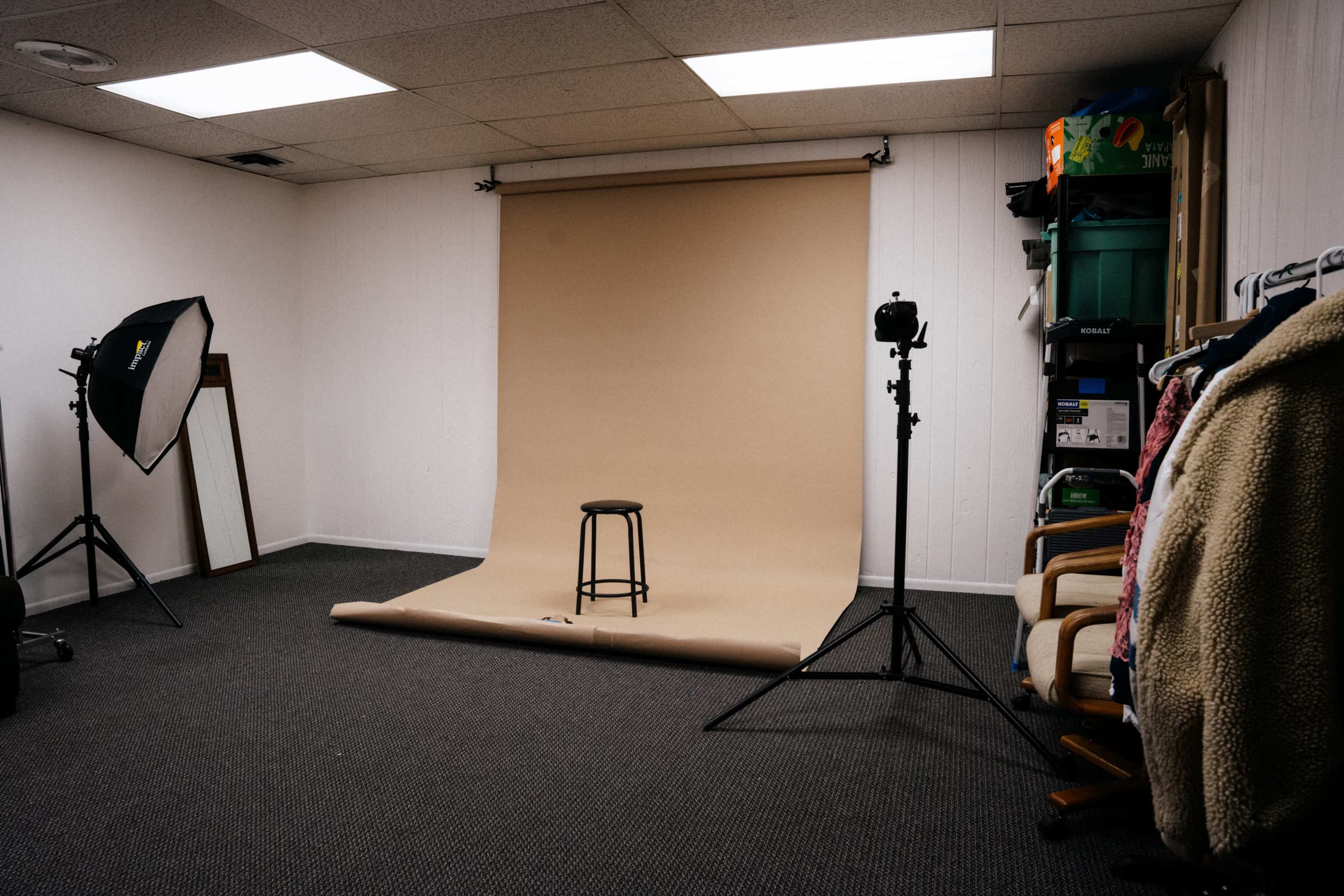 The image shows a photography studio with a beige backdrop, a stool in the center, and equipment including a lighting setup and a camera on a tripod.