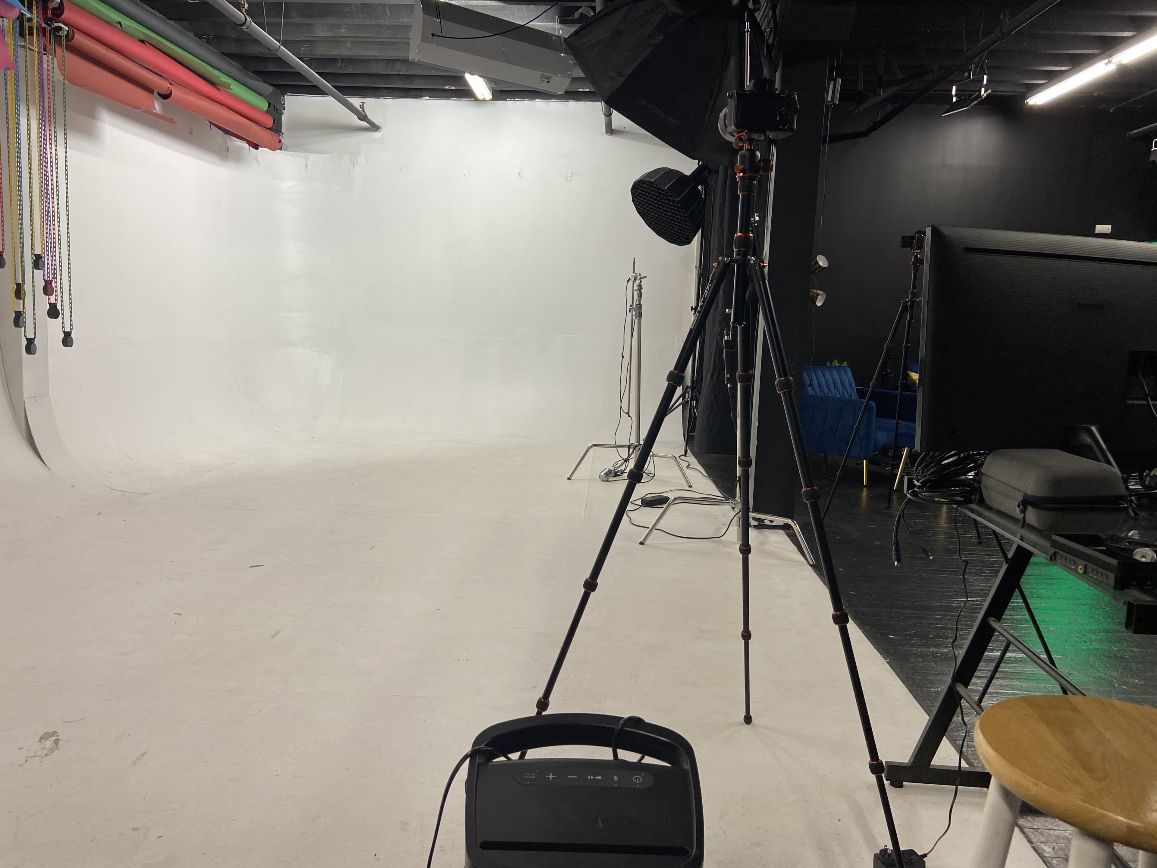 A photography studio with a white backdrop, equipment including a tripod, lighting setup, and a monitor.