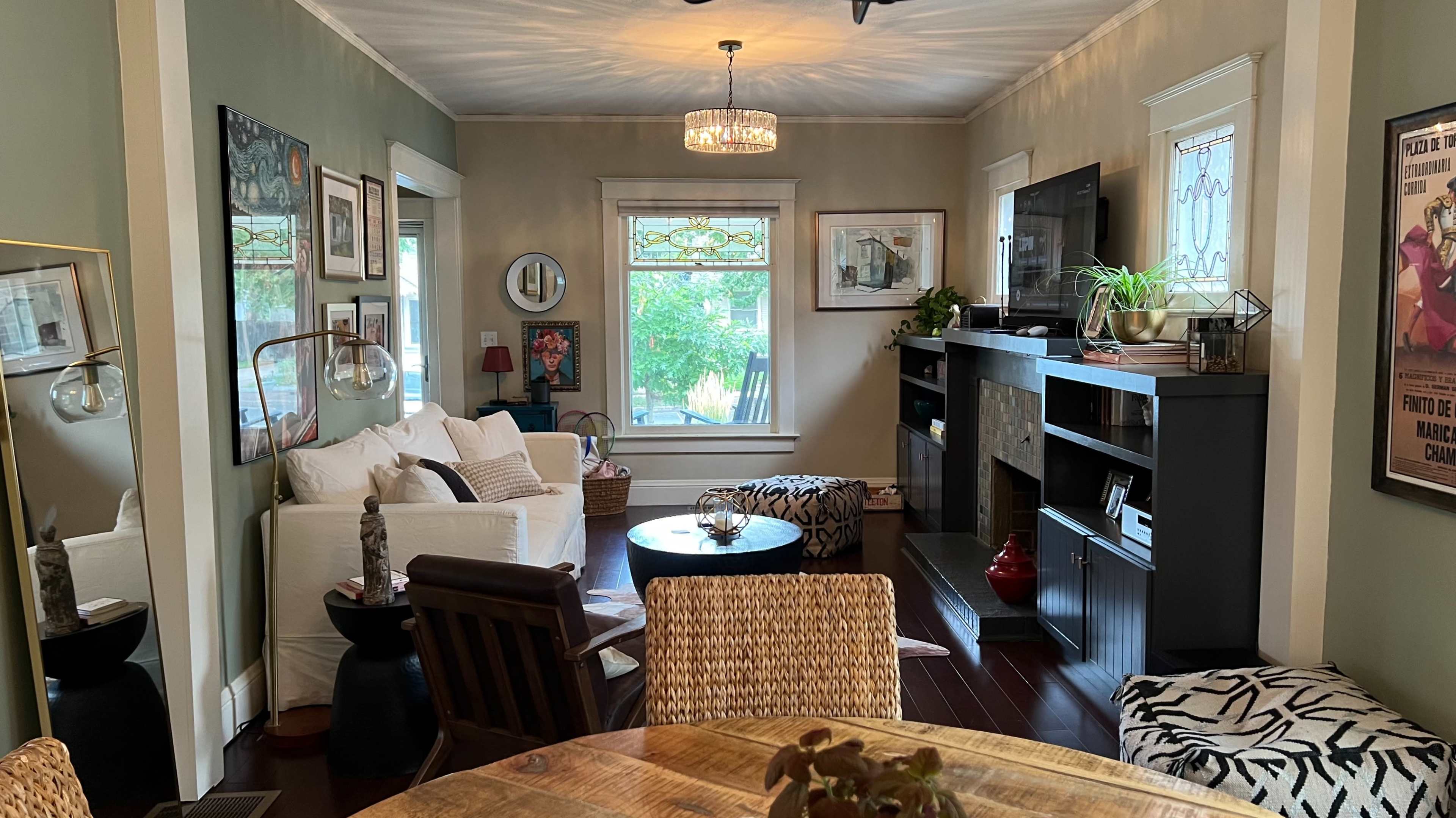 The image shows a cozy living room with a mix of seating areas, a dining table, and large windows allowing natural light to enter.