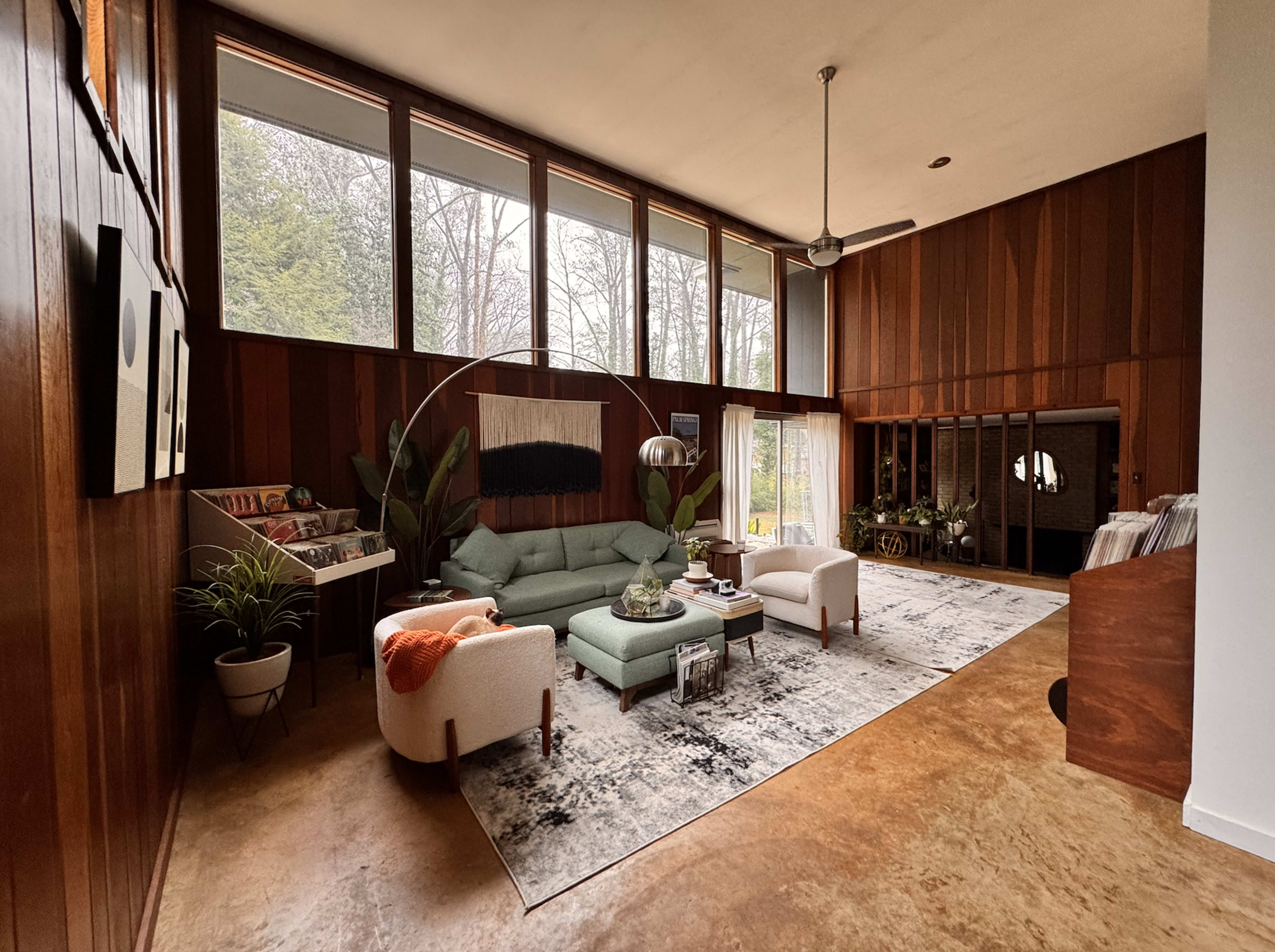 The image shows a spacious living room with large windows, wooden paneling, a green sectional sofa, and a decorative area rug.