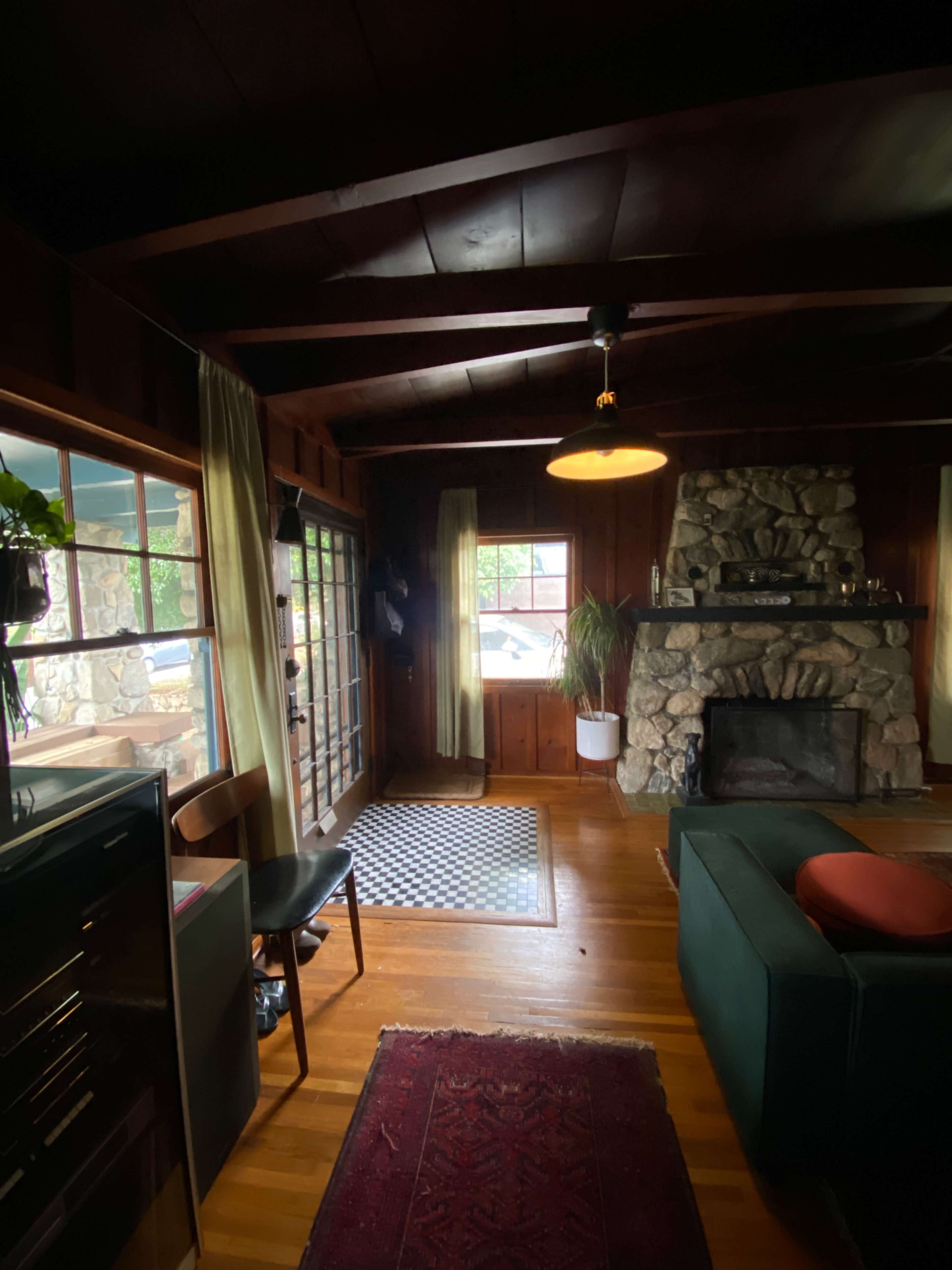 The image shows a cozy living room with a stone fireplace, wooden walls, and large windows allowing natural light to enter.