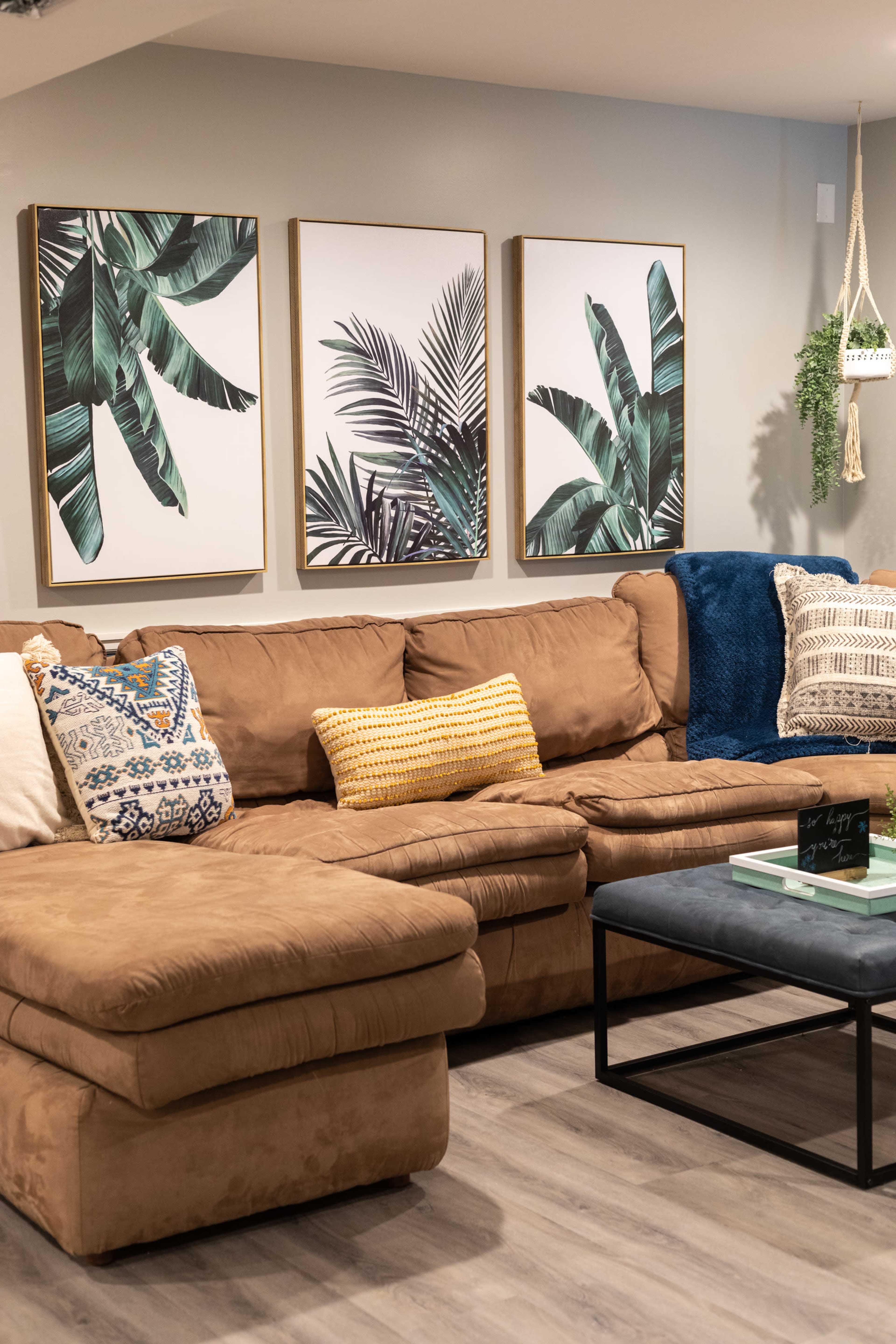A large brown sectional sofa is positioned in front of three framed botanical prints on a wall, with decorative pillows and a coffee table in the center.