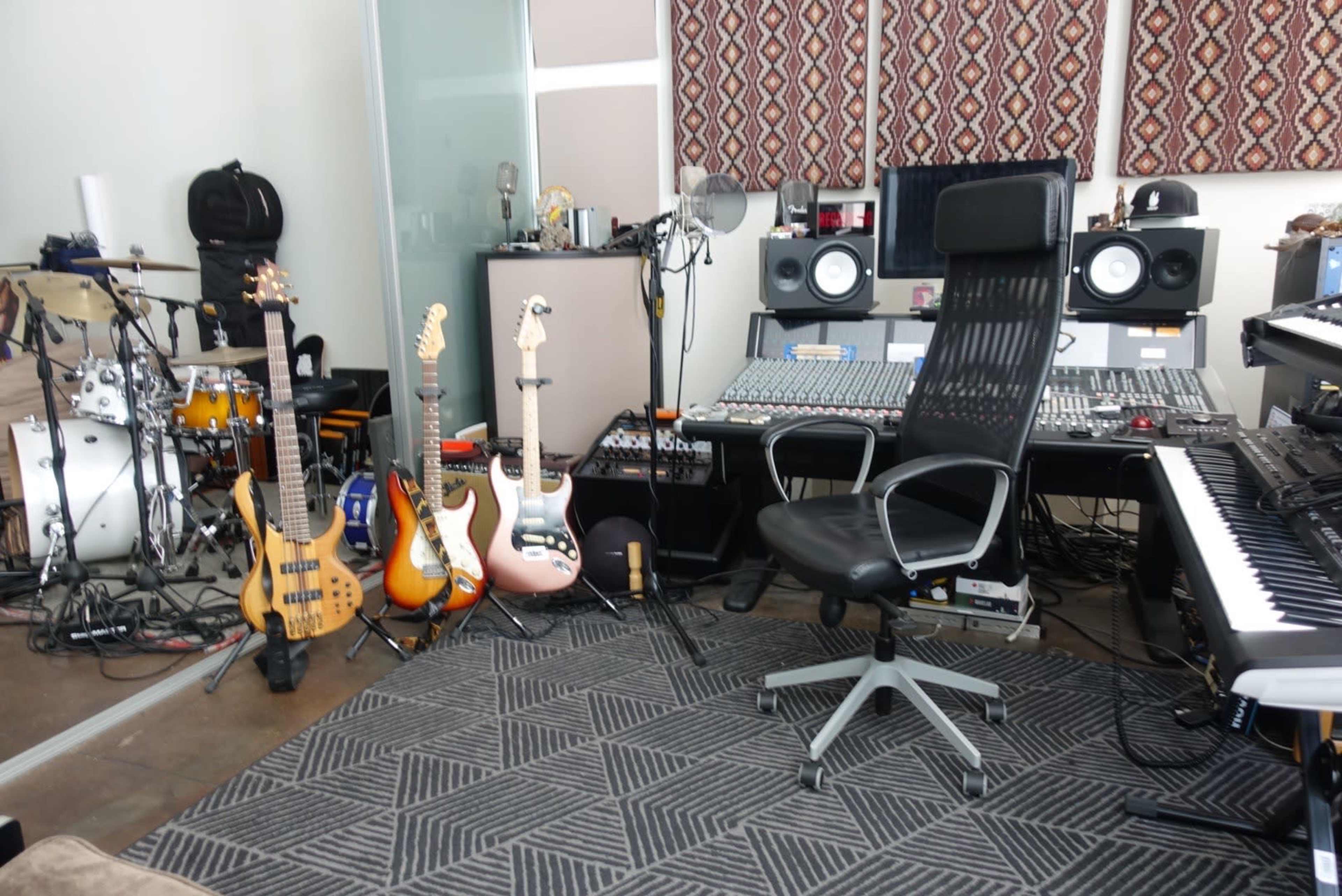 A music studio with various instruments, including guitars and drums, alongside a black office chair and a sound mixing console.
