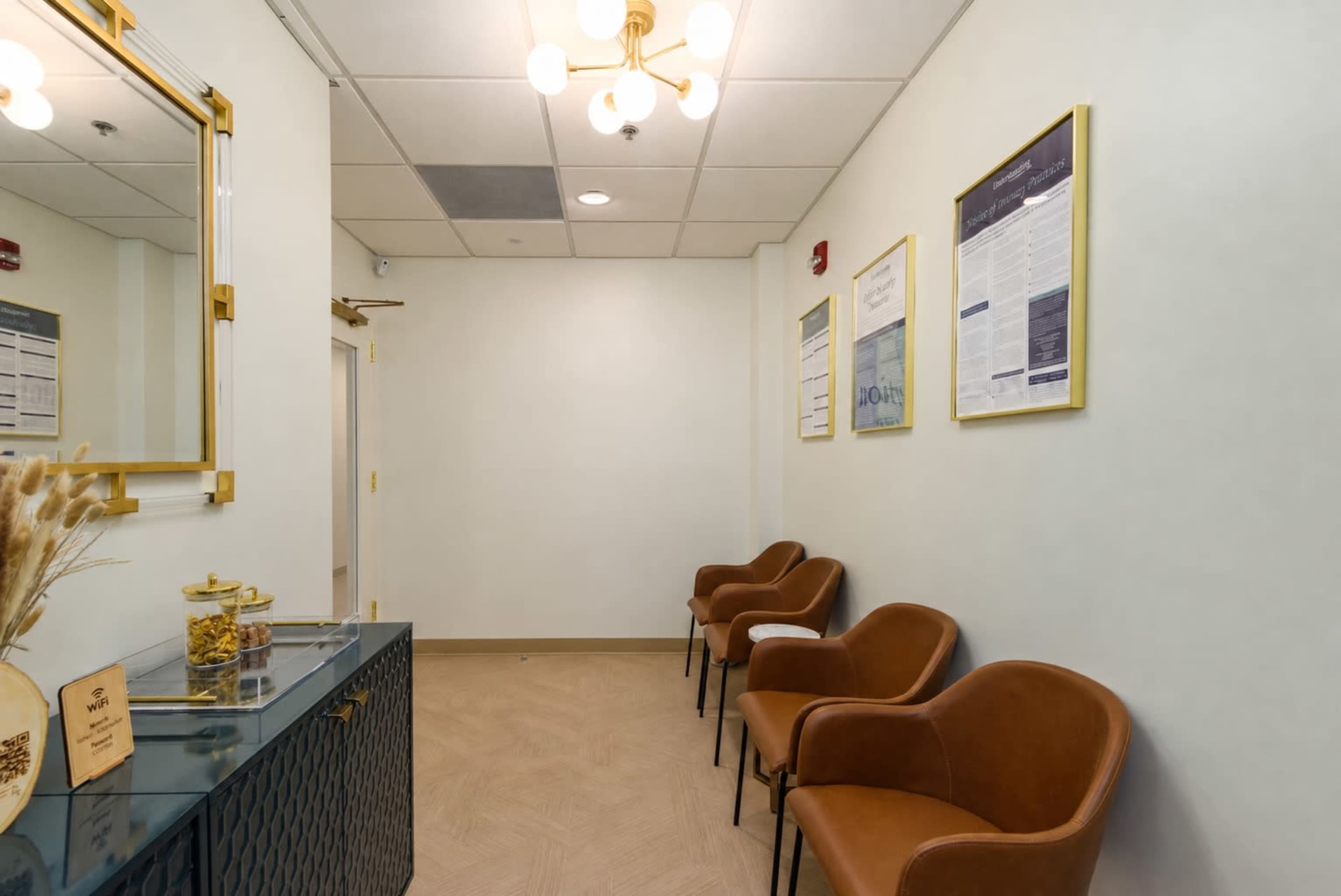 The image shows a waiting area featuring a small console table, three brown chairs, and informational posters on the walls.
