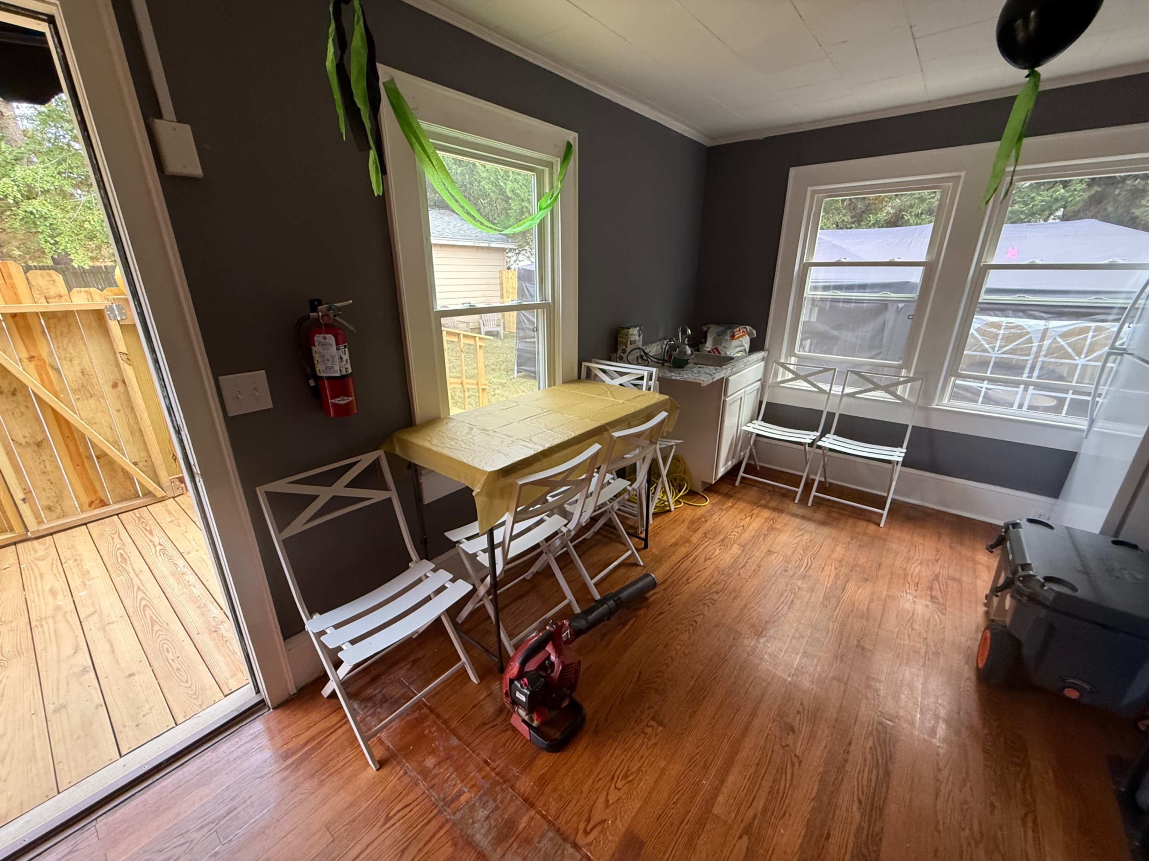 The image shows a small dining area with a yellow table and several white folding chairs, along with a wooden floor and a partially visible outdoor space.