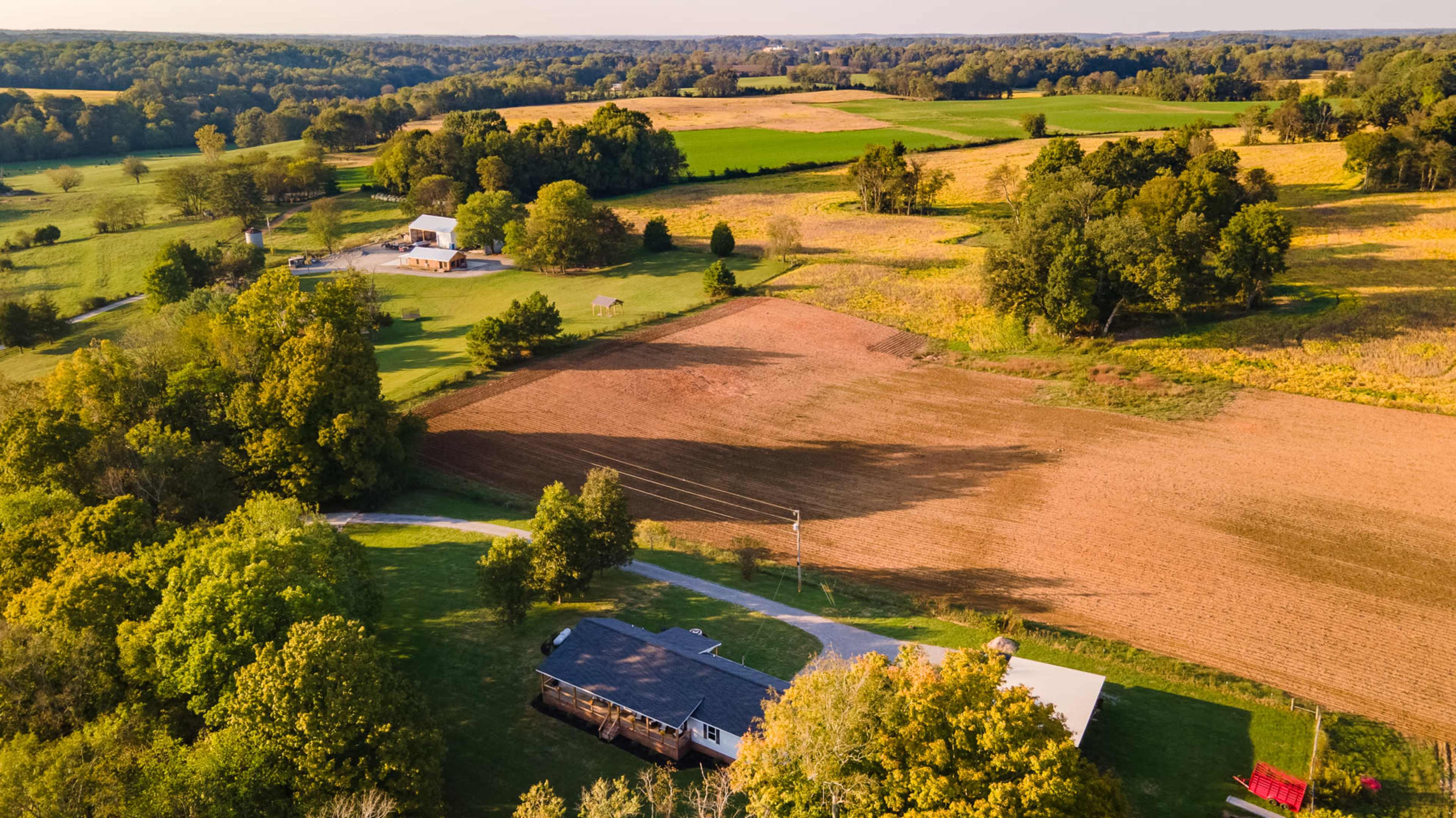 The aerial view shows a rural landscape with farmland, a house, fields, and patches of trees under a clear sky.