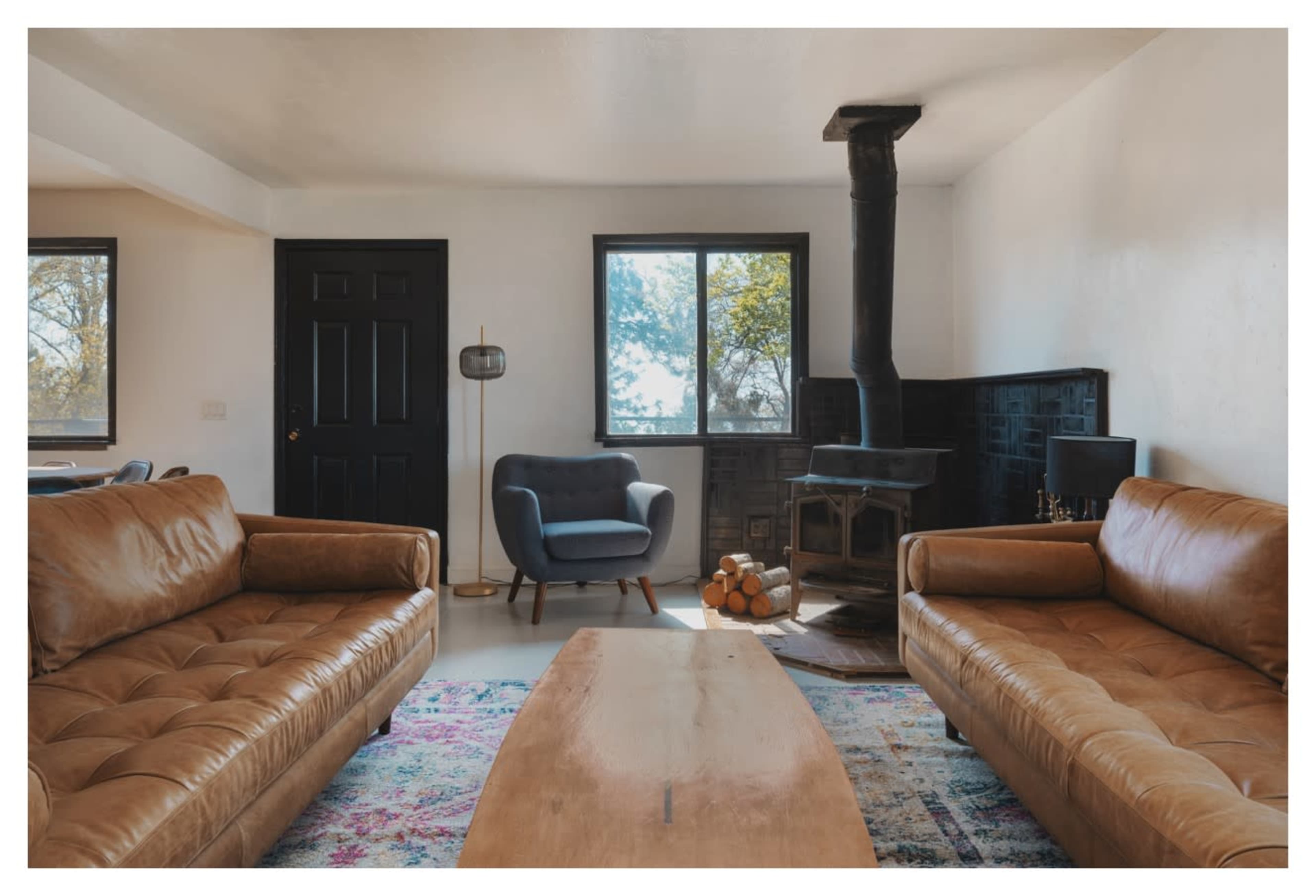A modern living room with two leather sofas, a wooden coffee table, a wood stove, and a large window allowing natural light to enter.