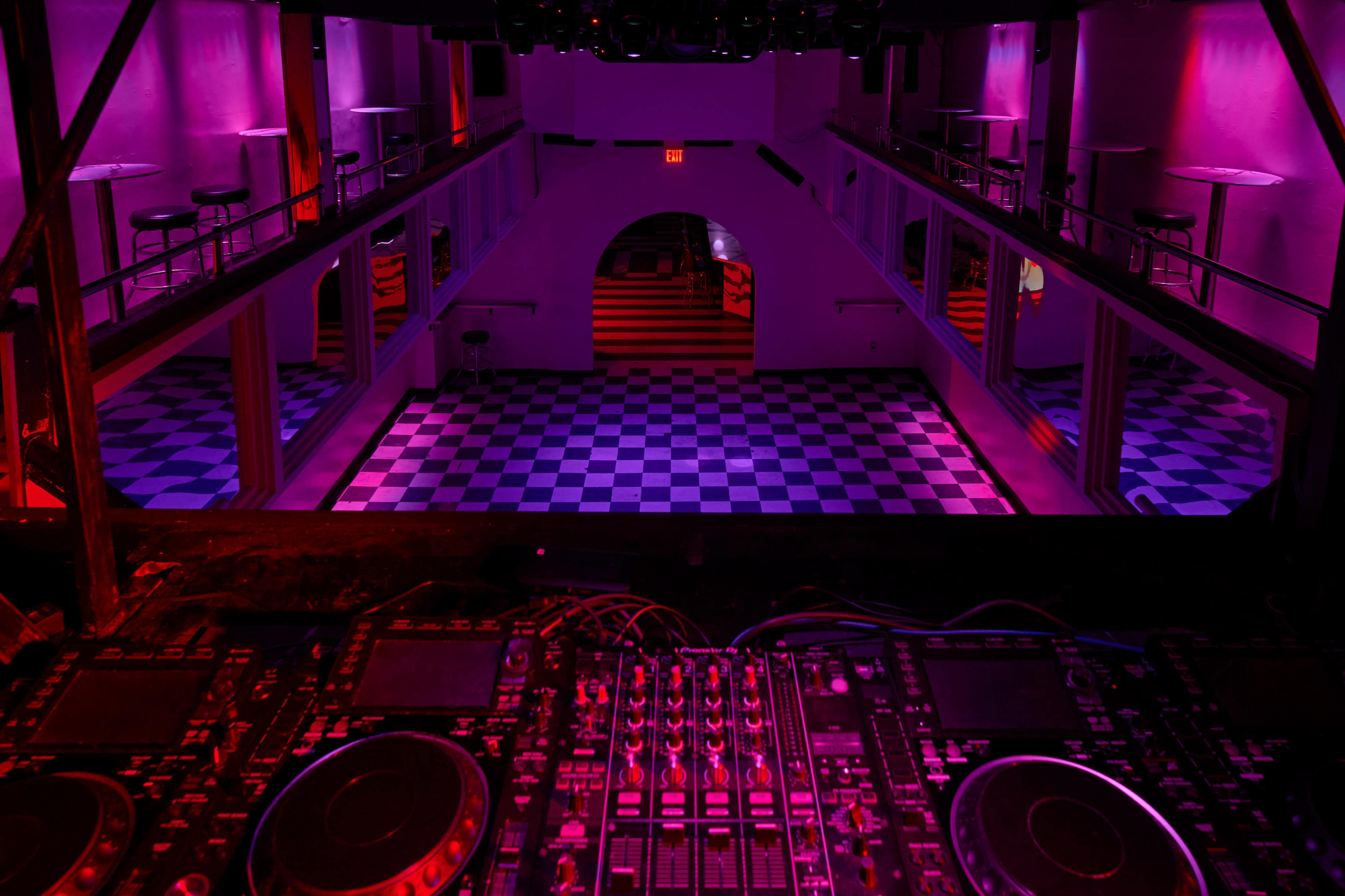 An empty nightclub from a high vantage point, featuring a checkered dance floor and illuminated walls in shades of pink and purple.