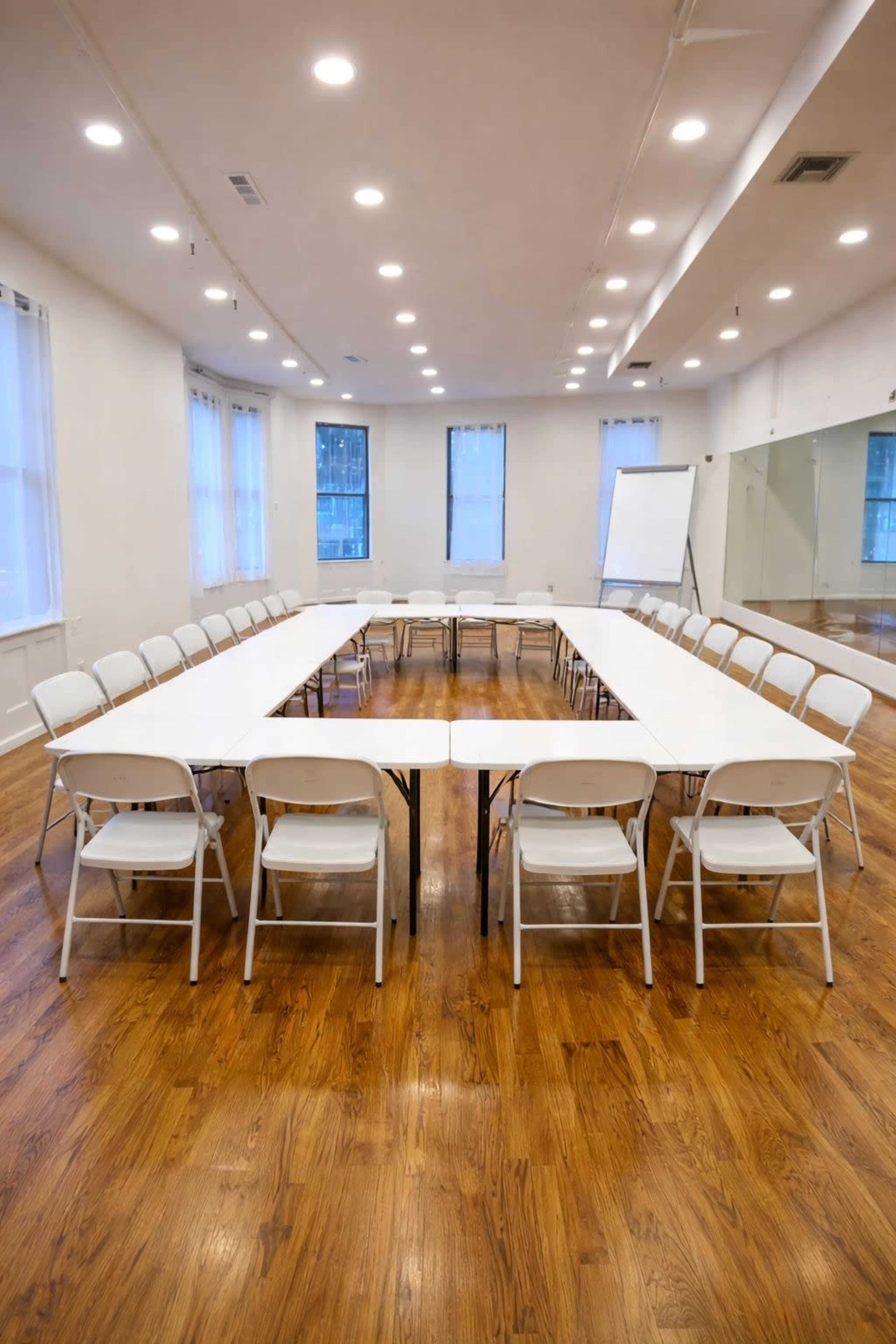 A large meeting room features a U-shaped arrangement of white folding chairs around a long table, with hardwood floors and natural light from nearby windows.