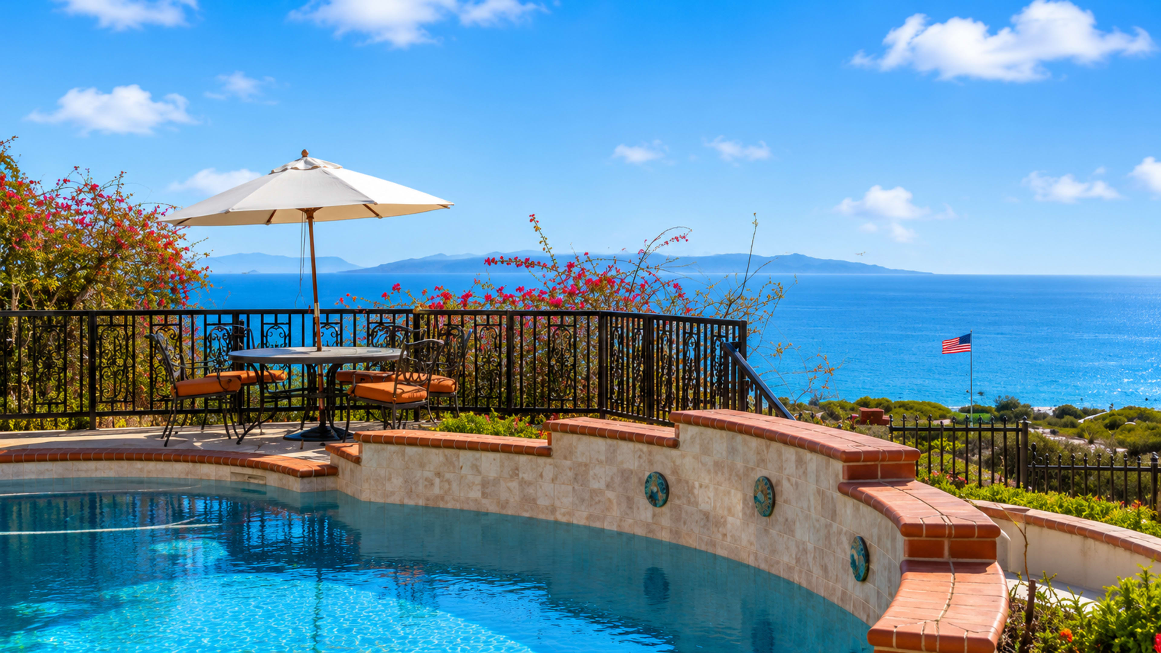The scene features a swimming pool surrounded by vibrant flowers, with a patio table and umbrella overlooking the ocean and distant islands.