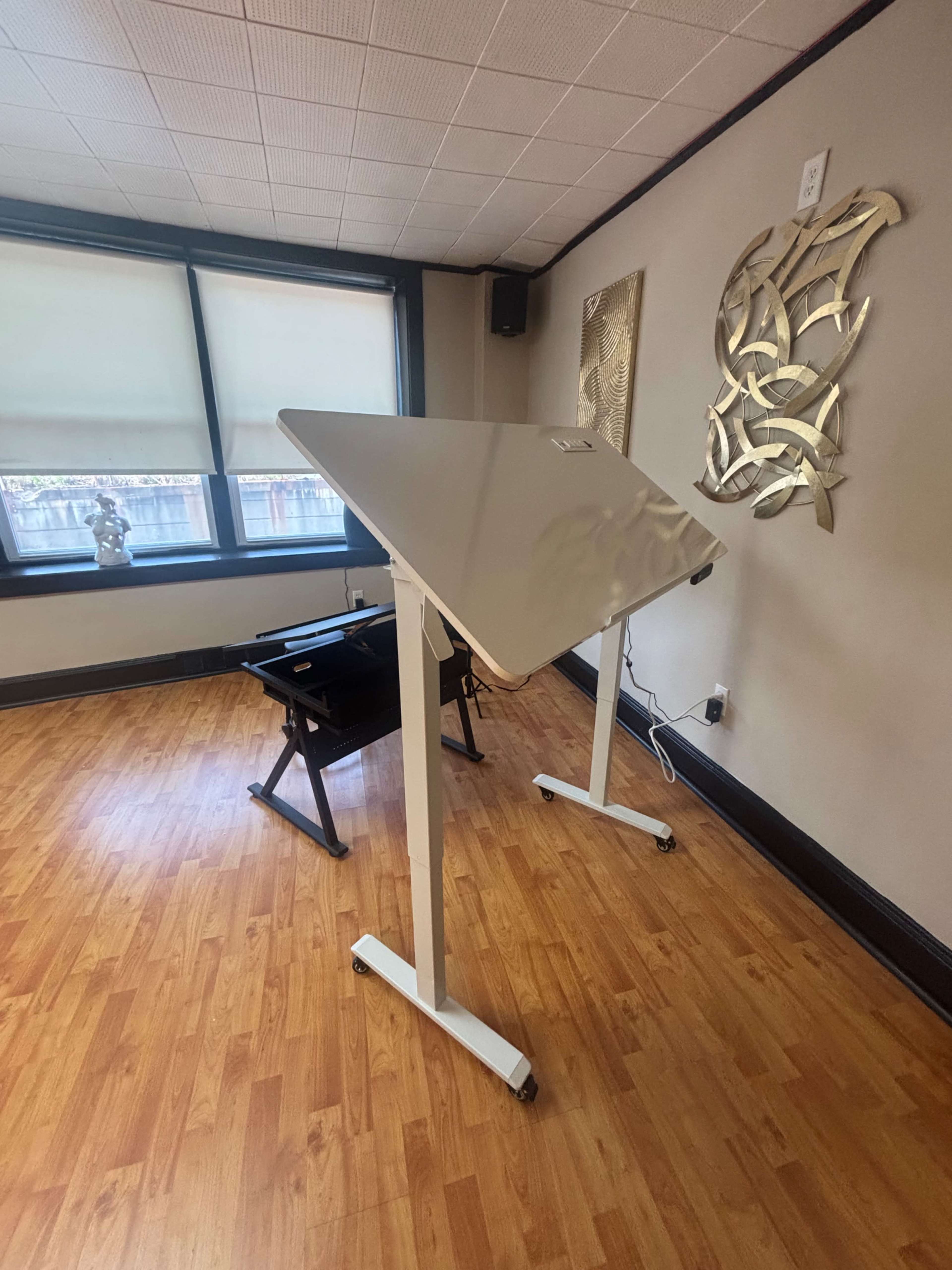 A modern adjustable desk is positioned in a well-lit room with wooden flooring and decorative wall art.