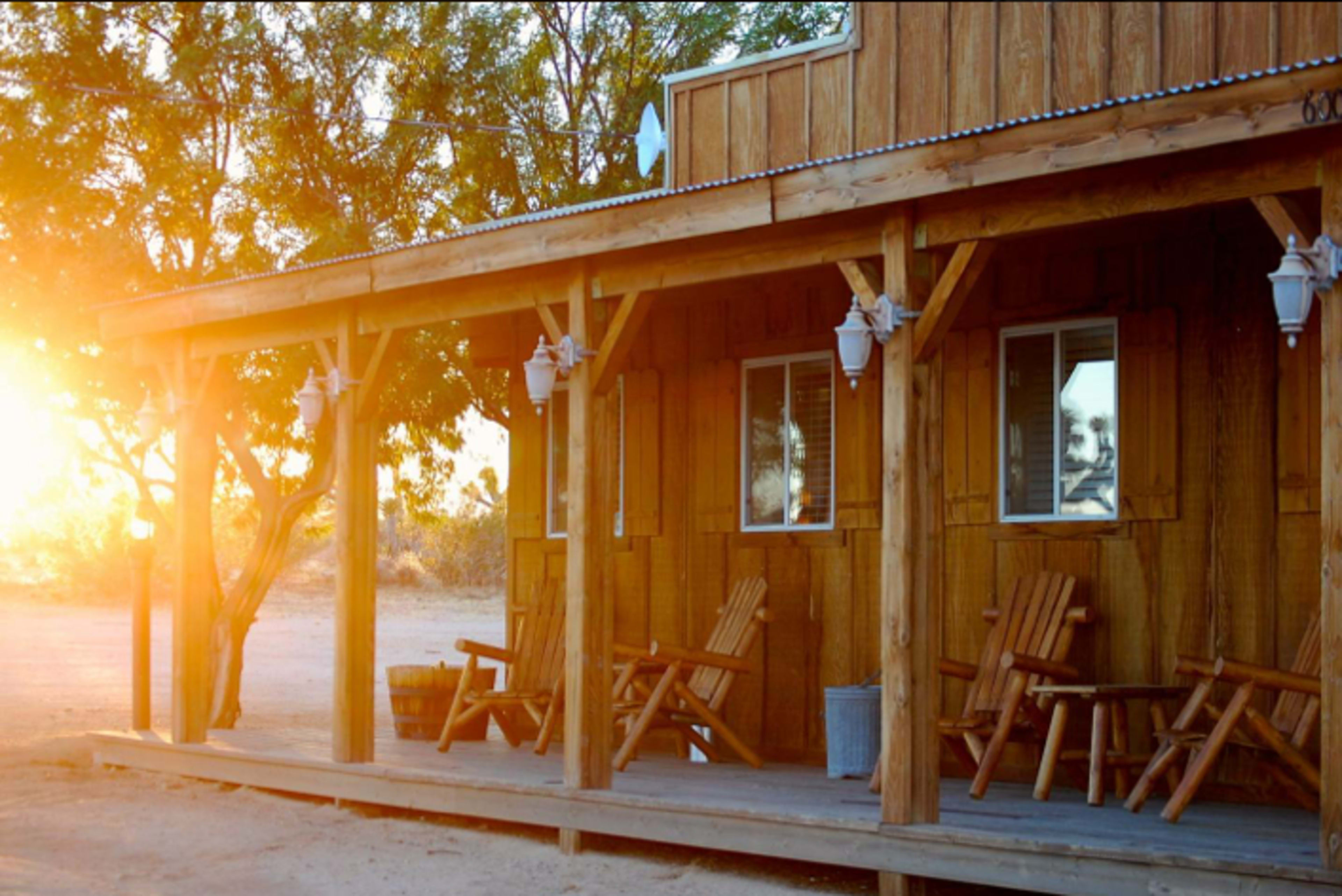 The image shows a wooden porch with several adirondack chairs and a cabin-like structure bathed in warm sunset light.