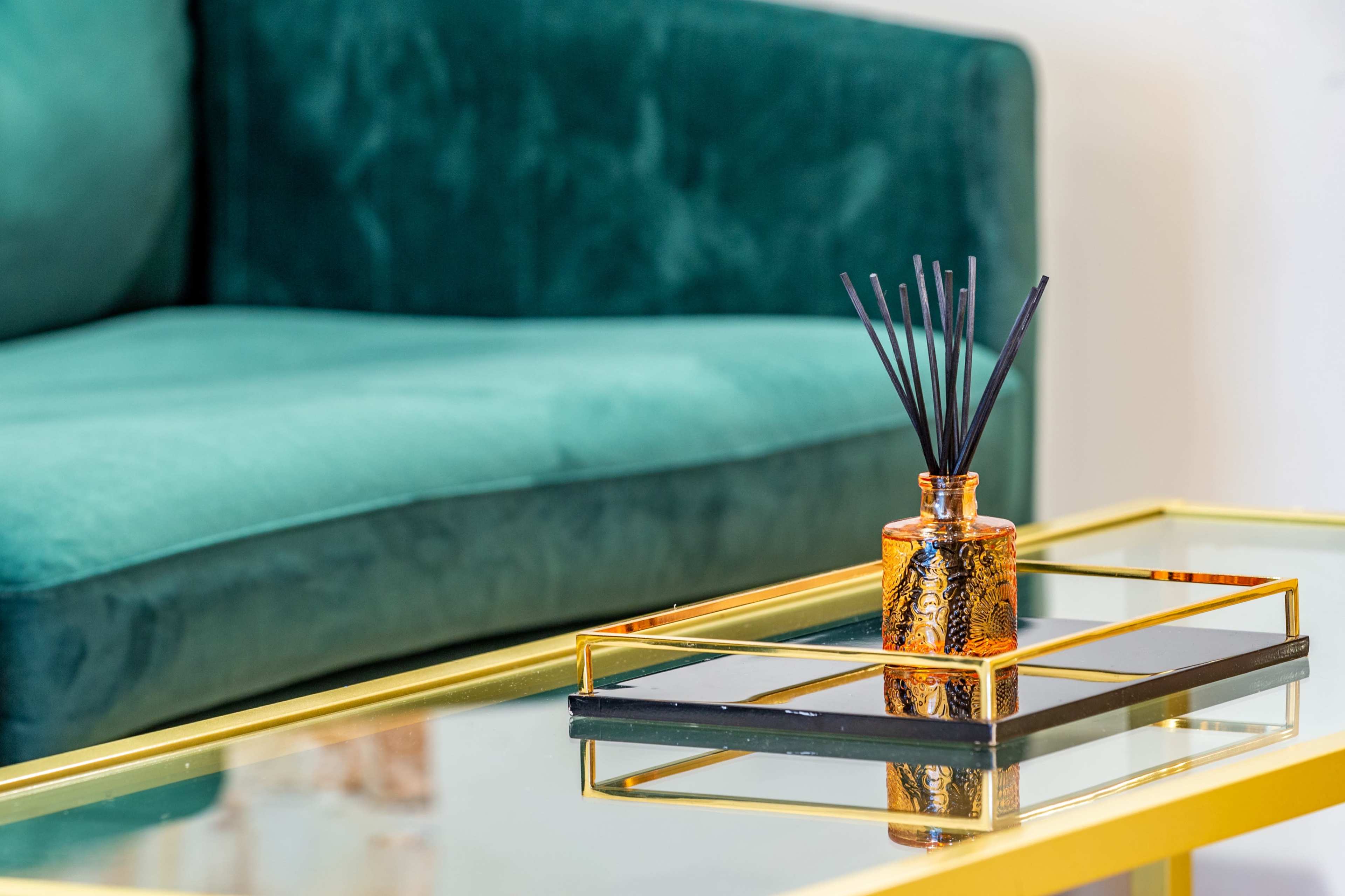A glass table with a gold frame and a black tray holds a small decorative bottle filled with reed sticks, positioned in front of a green velvet couch.