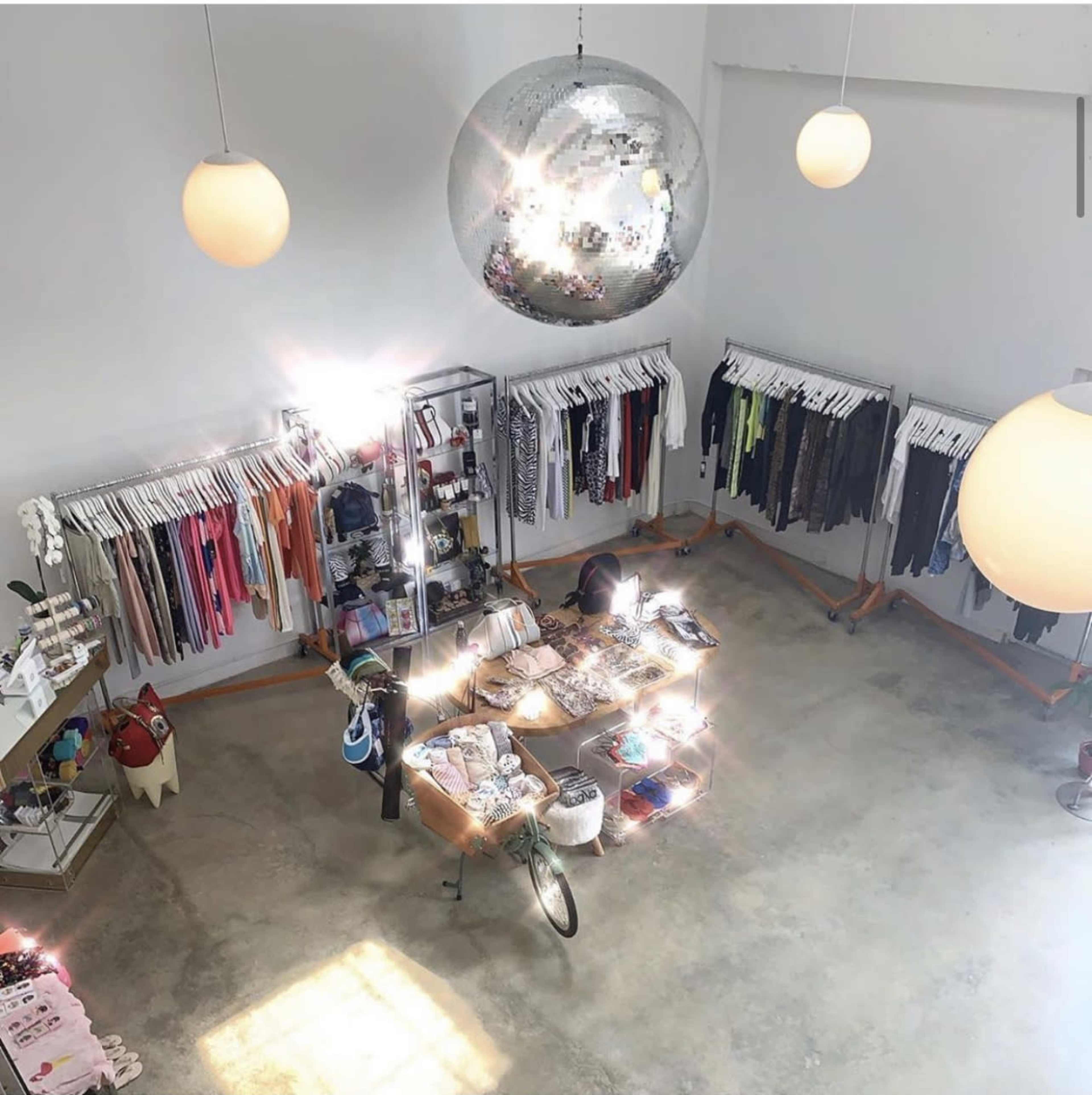 The image shows an aerial view of a clothing store with neatly organized apparel on racks and a central display table surrounded by large hanging light fixtures.