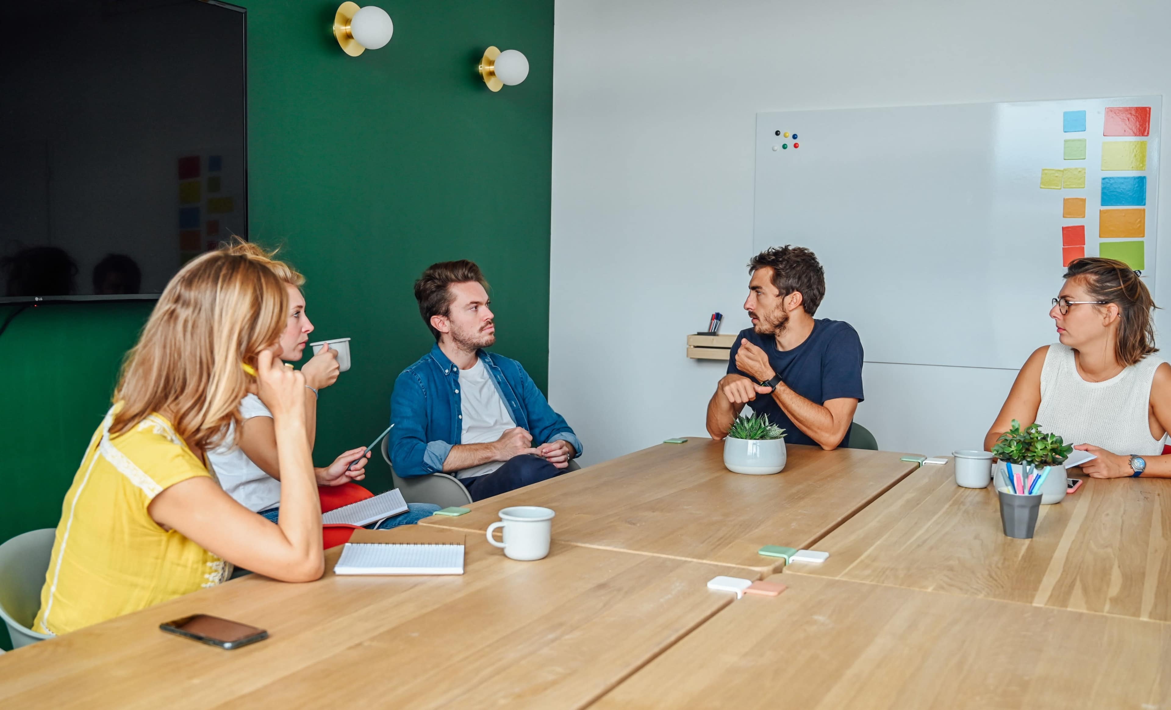 A group of five people engage in a discussion around a wooden table in a well-lit conference room with green accents and a whiteboard.