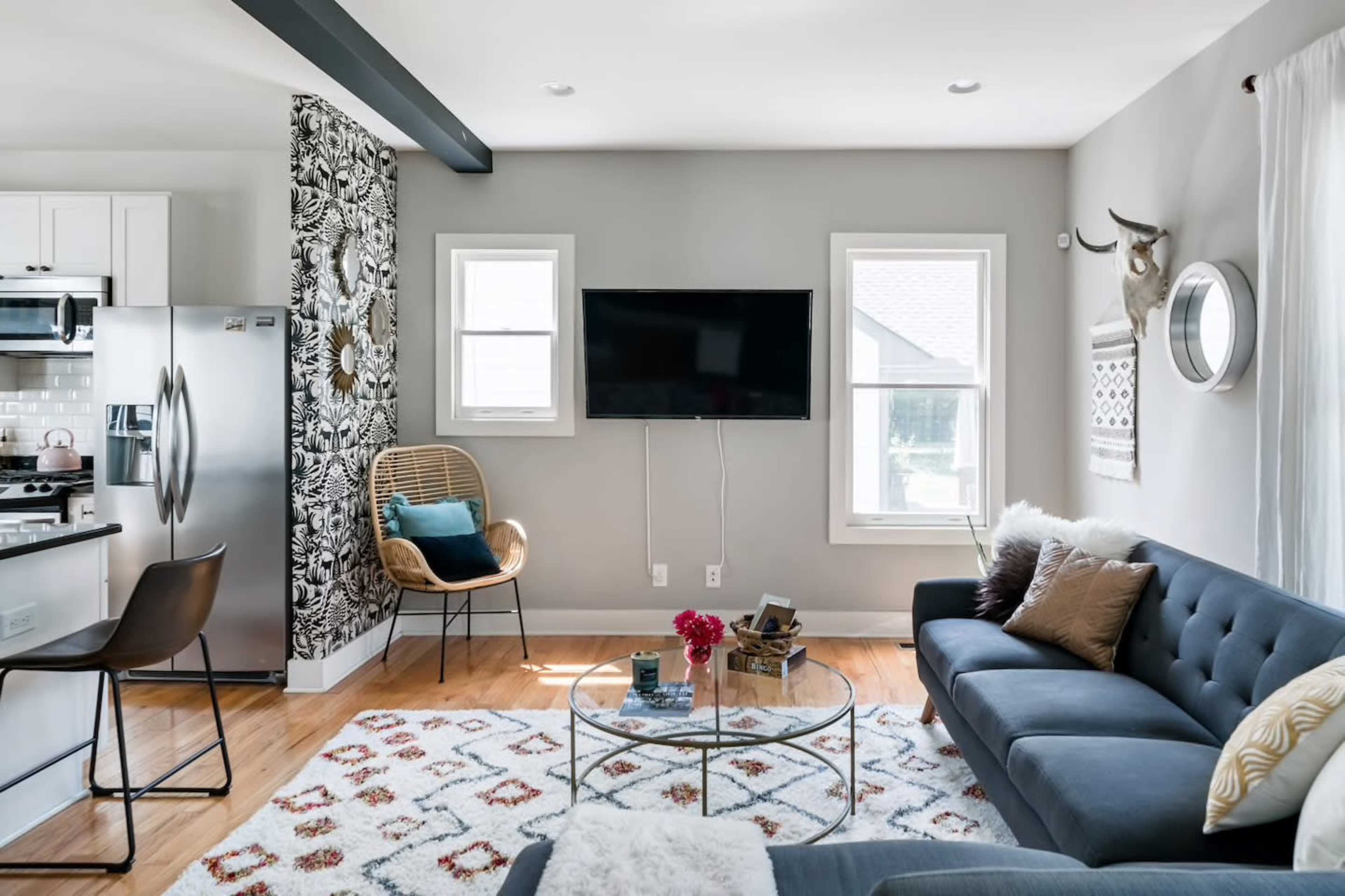 A modern living room features a gray sofa, a glass coffee table, a television mounted on the wall, and a patterned area rug.