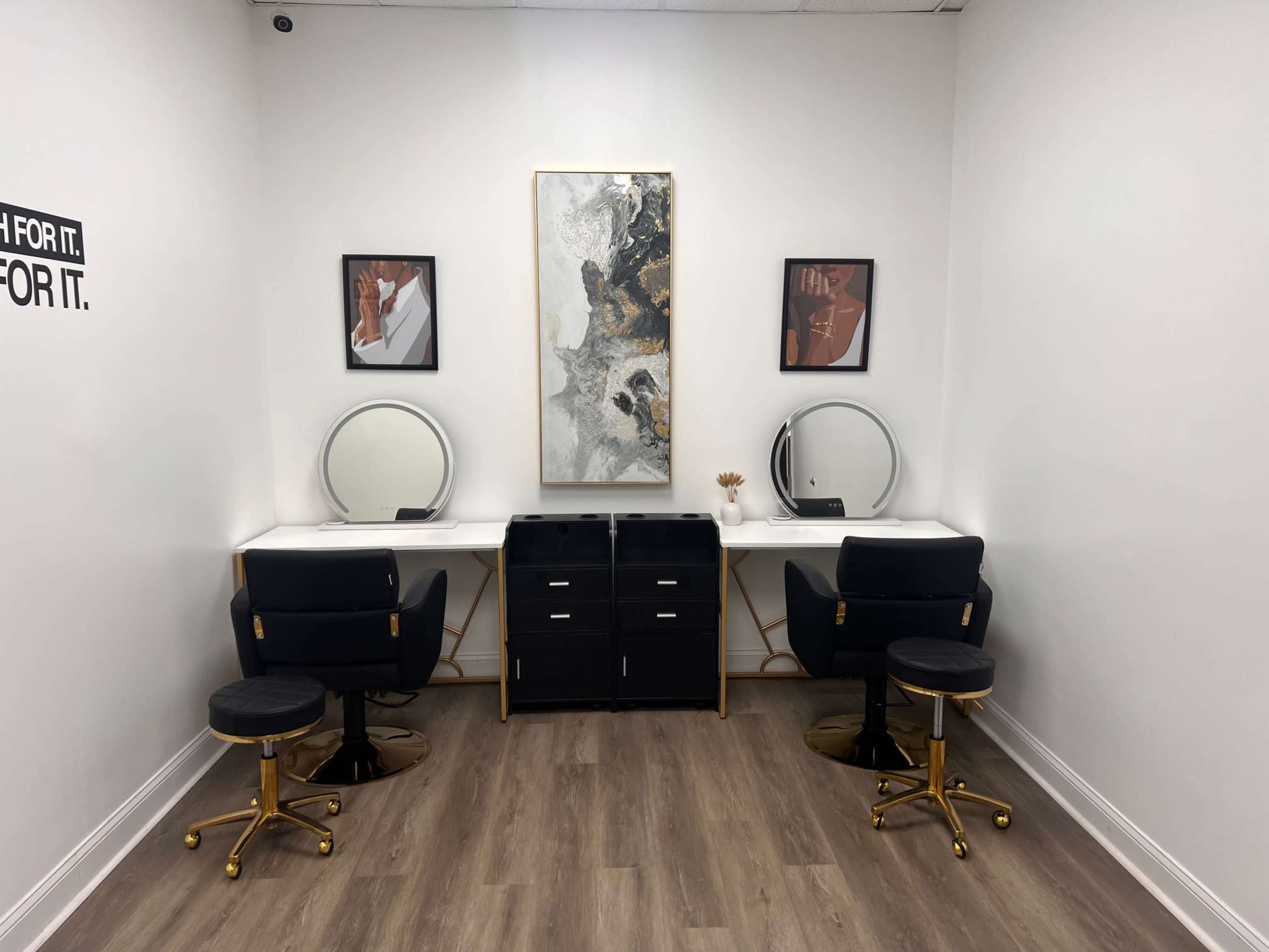 The image shows a well-lit salon room featuring two vanity stations with circular mirrors, black and gold seating, and a modern piece of wall art between two framed pictures.