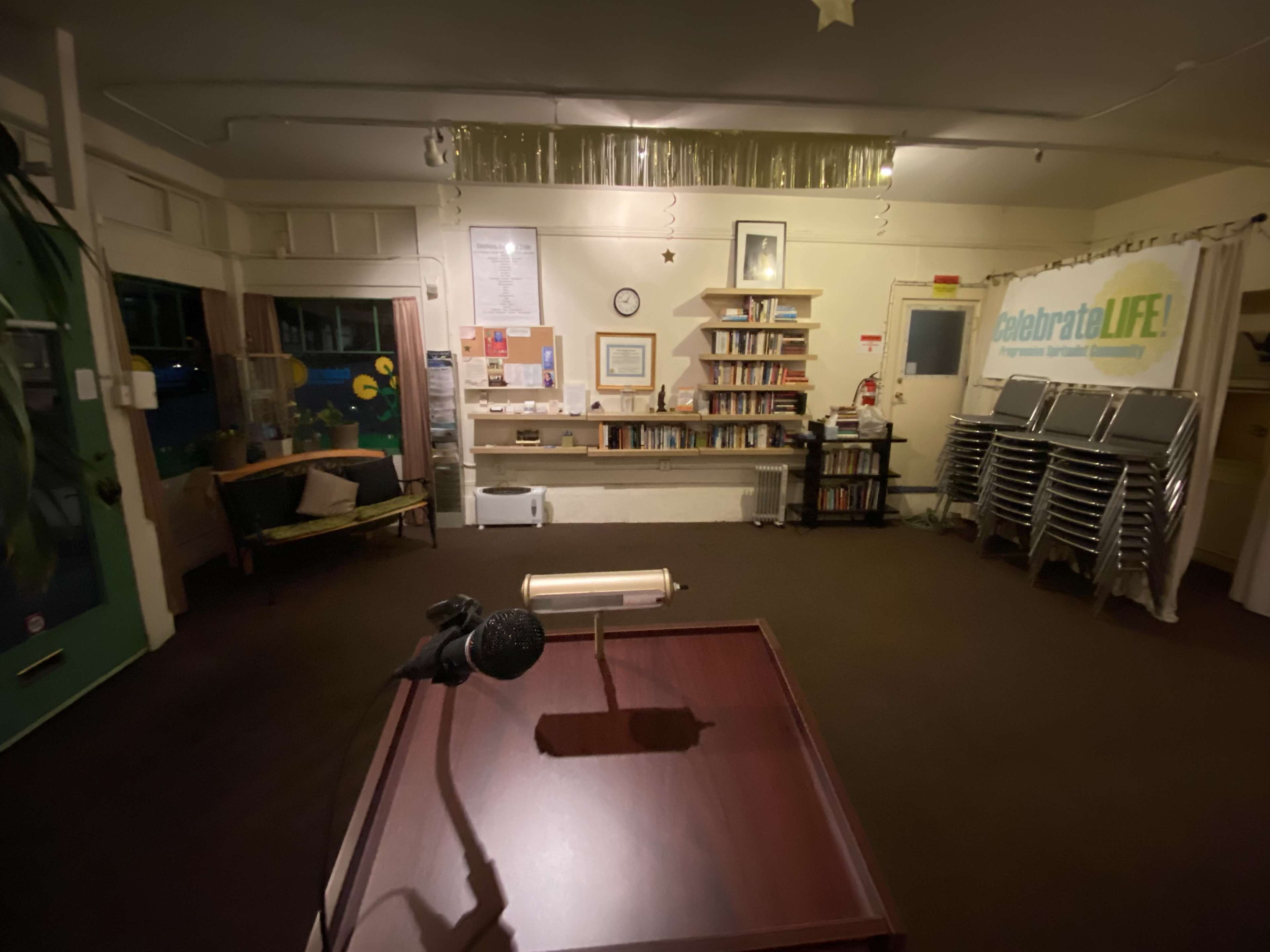 The image shows a sparsely furnished room with a podium and microphone in front, bookshelves along the back wall, and stacked chairs to the right.