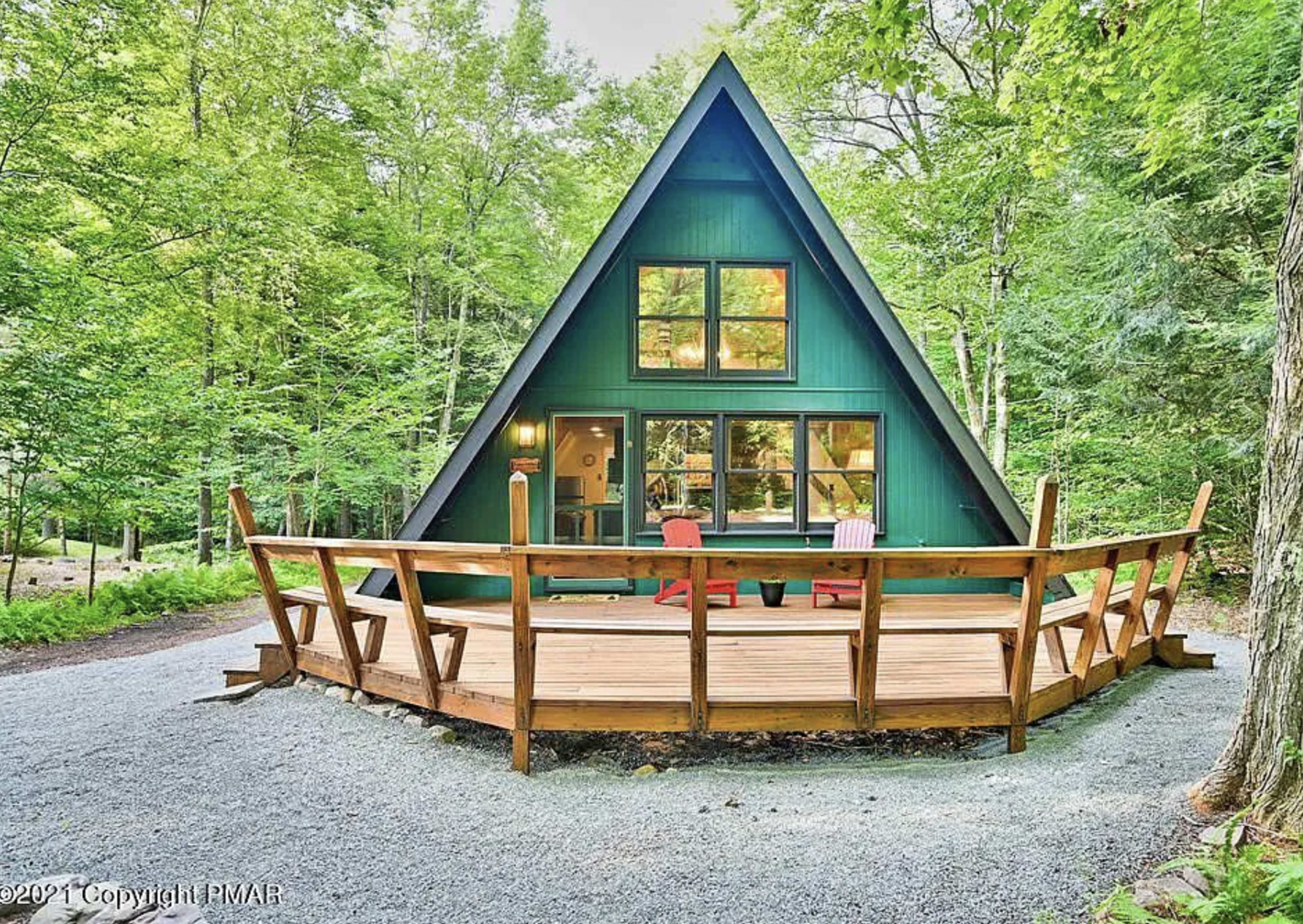 A blue A-frame house with large windows is surrounded by trees and features a wooden deck with chairs in front.