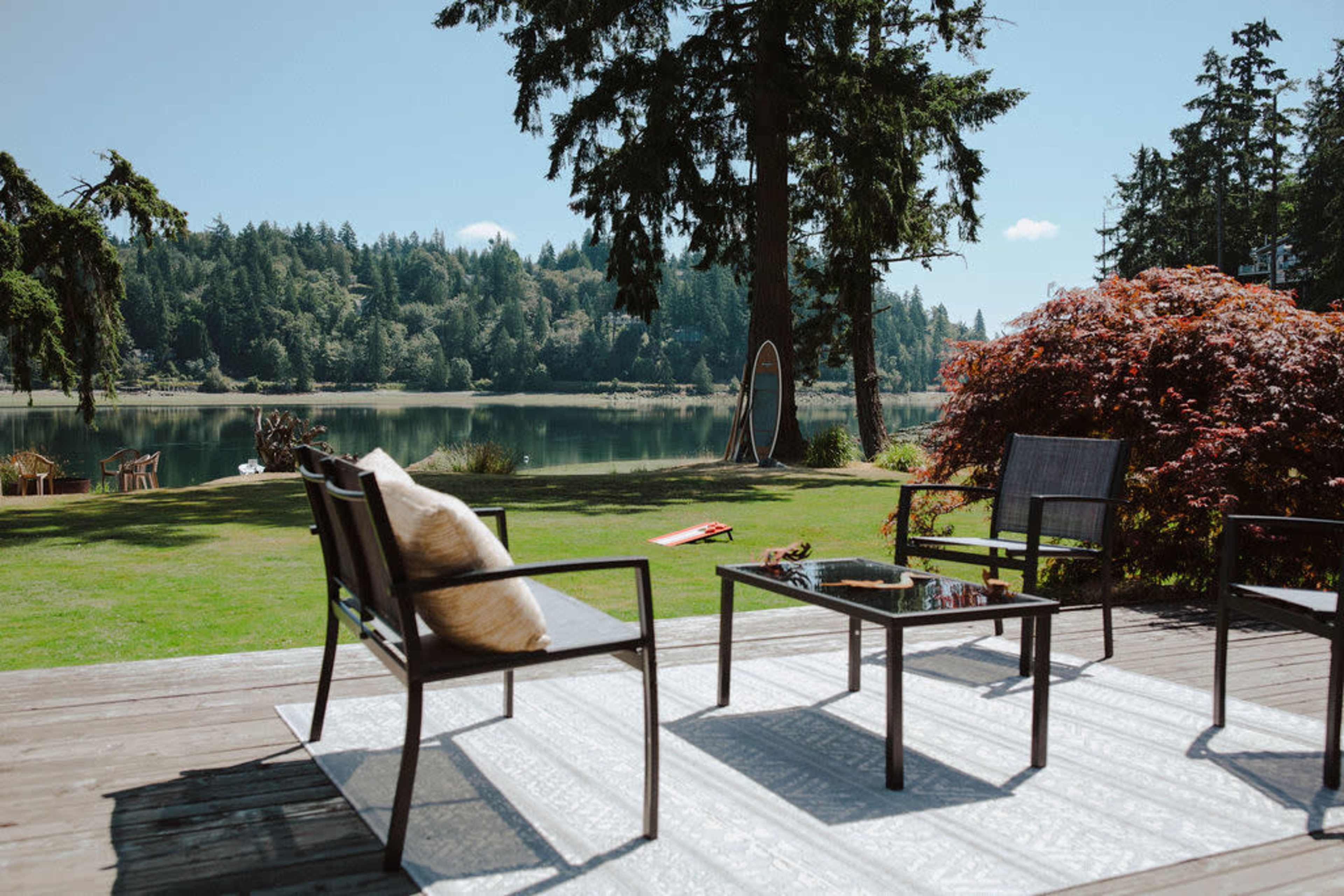 A wood deck features a set of black chairs and a table, overlooking a calm lake surrounded by trees and a colorful shrub.