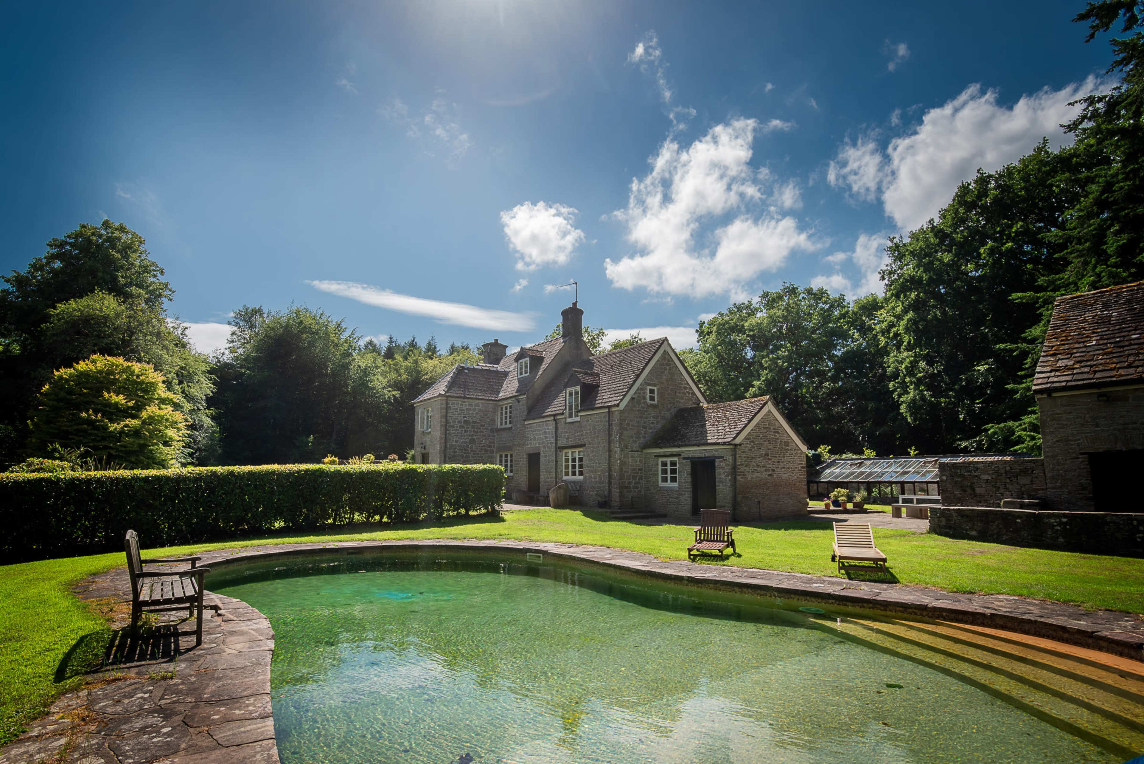 A stone house with multiple gables is surrounded by greenery and features a swimming pool in the foreground under a clear blue sky.