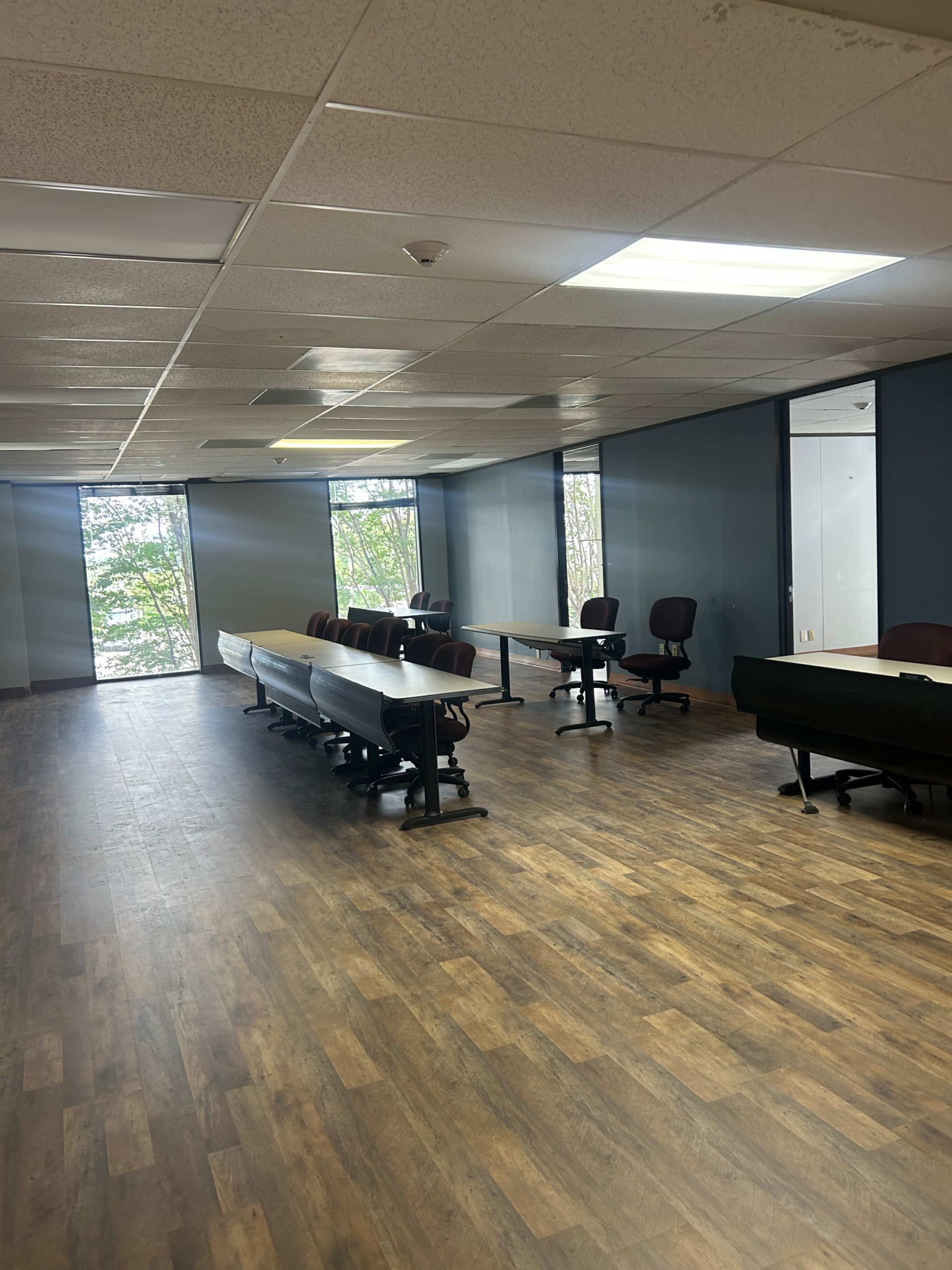 An empty office space features several tables and chairs arranged throughout, with large windows allowing natural light to enter.