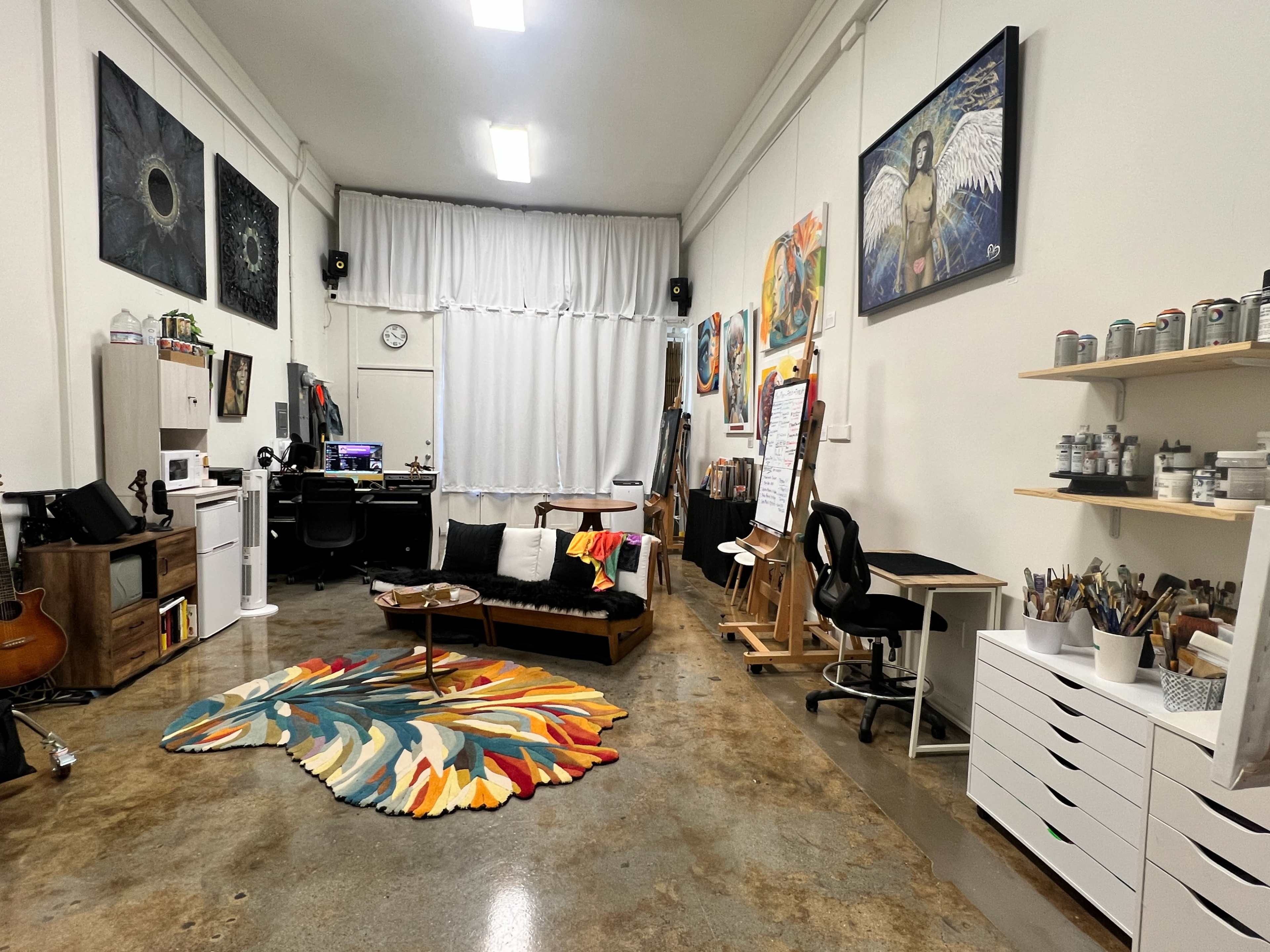 The image shows an art studio with various artworks displayed on the walls, a seating area with a colorful rug, and supplies organized on shelves.