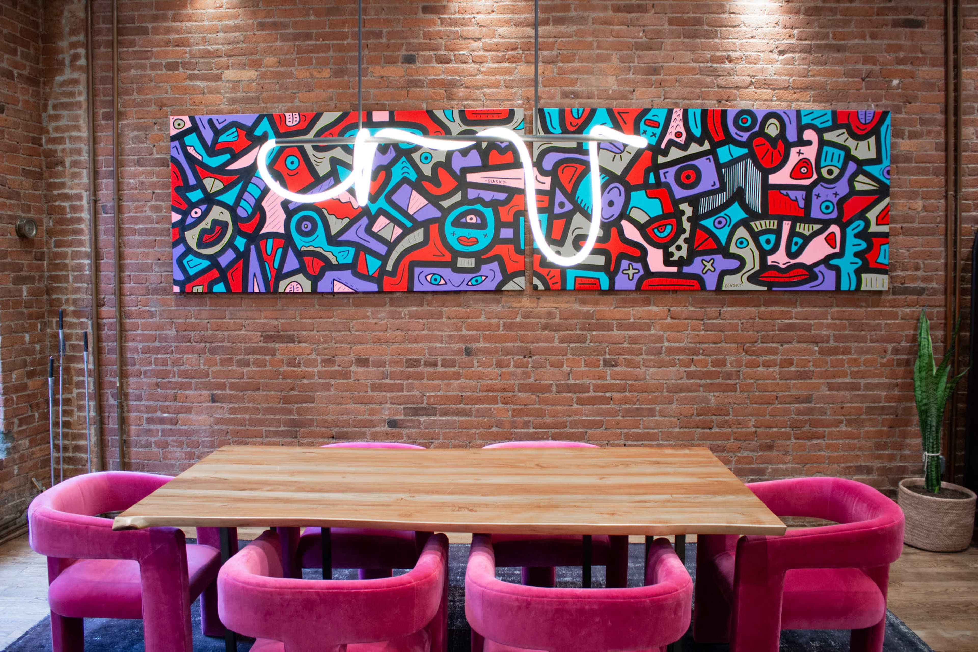 A large wooden table is set beneath a colorful abstract mural on a brick wall, surrounded by pink upholstered chairs.