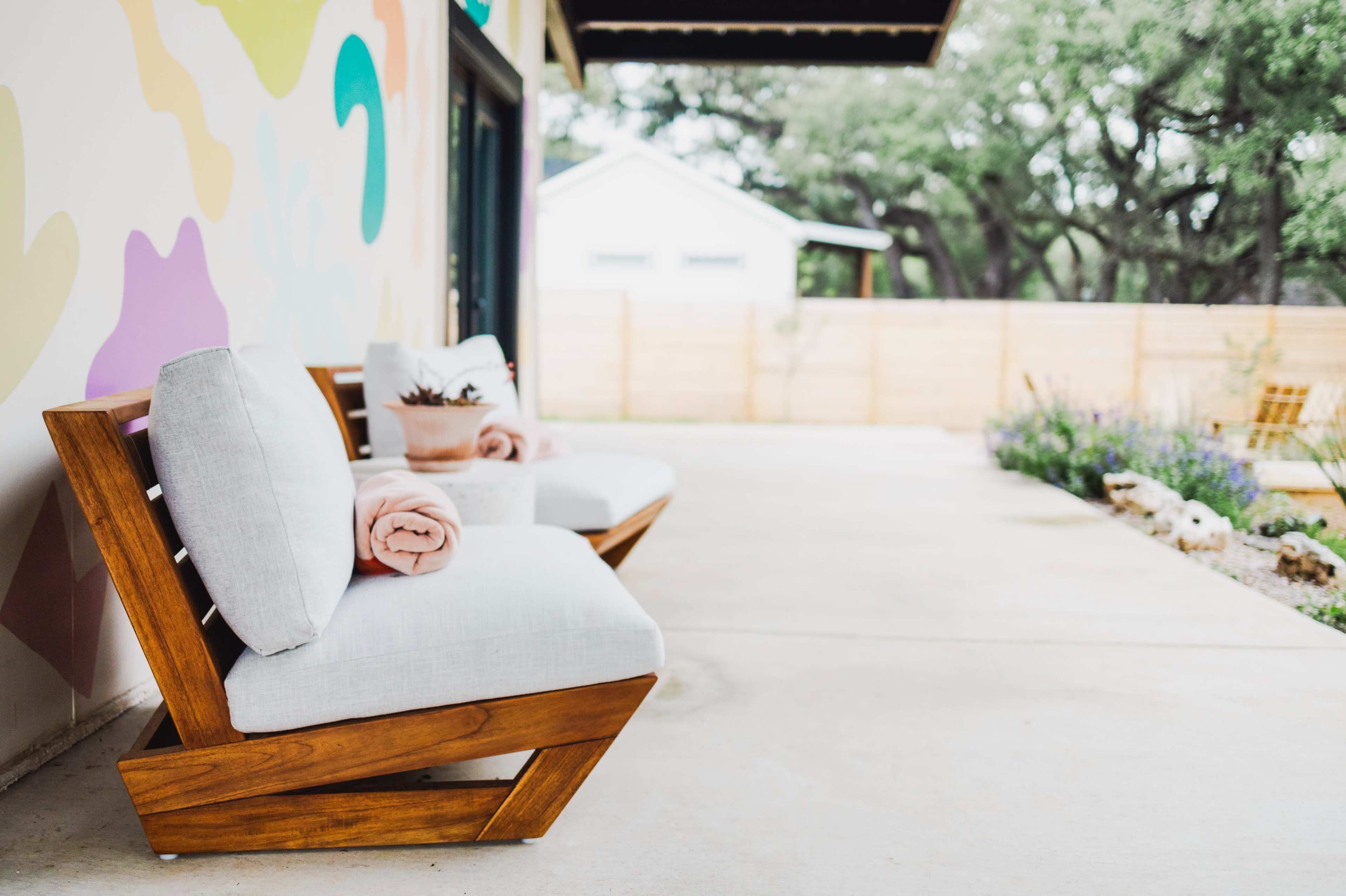 A concrete patio features two wooden chairs with light gray cushions, alongside a potted plant, in front of a colorful mural and a blurred backyard.