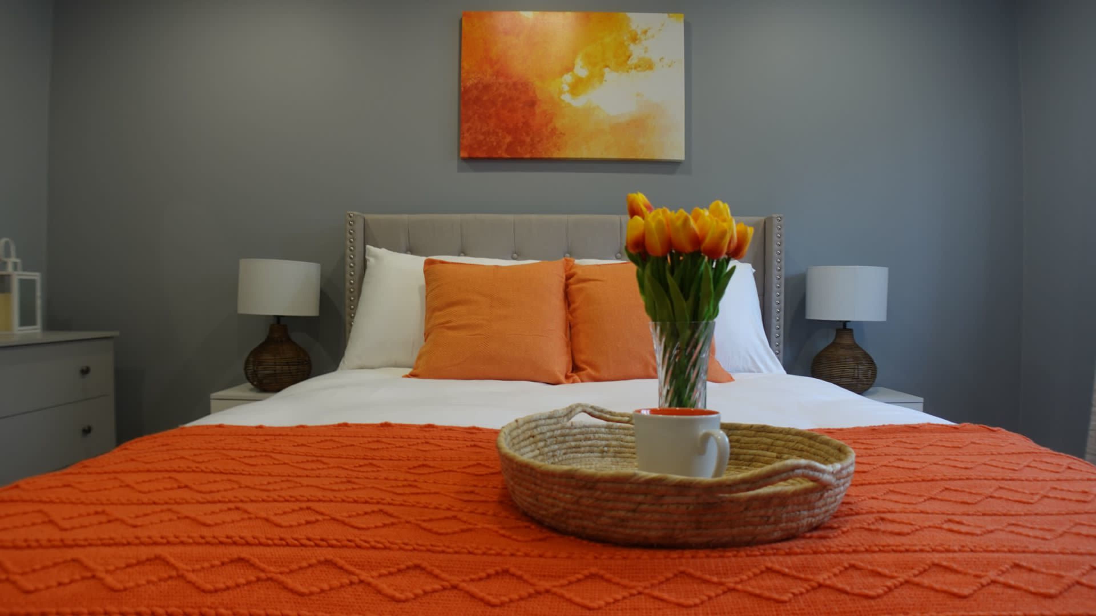 The image shows a neatly arranged bedroom with a large bed covered in an orange throw, flanked by two bedside lamps, and a vase of tulips on a tray in the foreground.