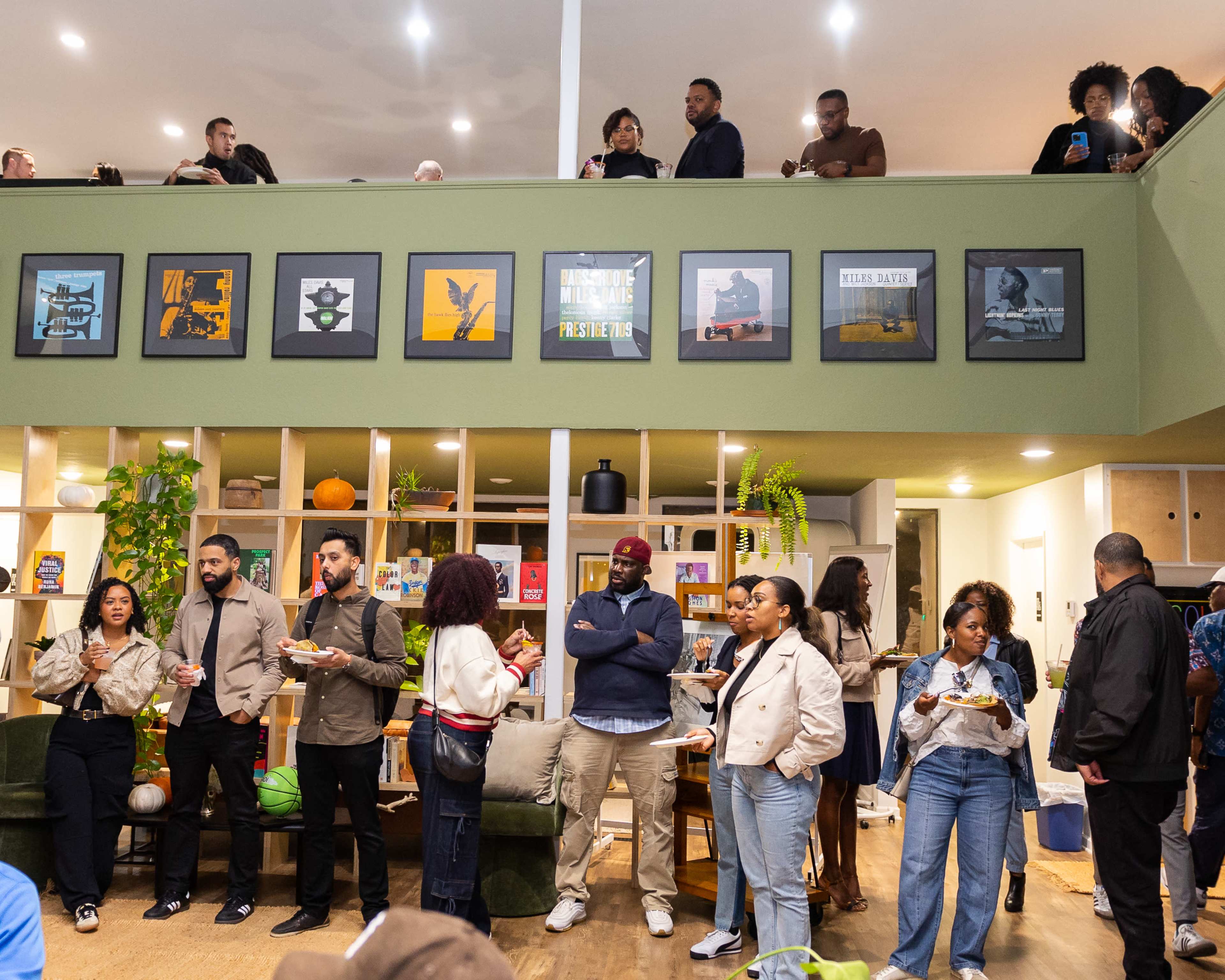 A diverse group of people socialize and enjoy food in a lively indoor space with green walls and framed artwork.