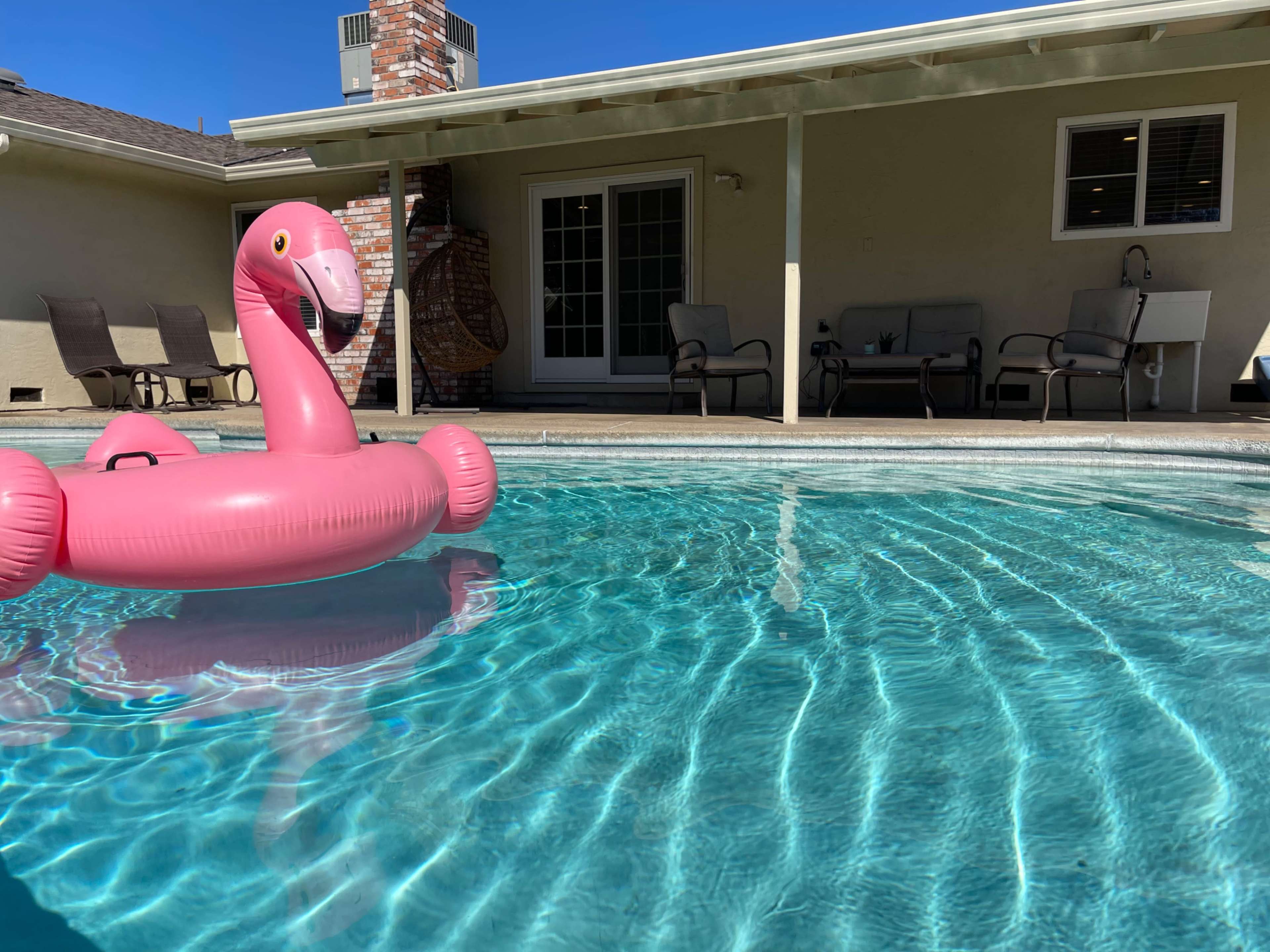 A pink inflatable flamingo floats in a clear blue swimming pool in front of a house with a patio and chairs.