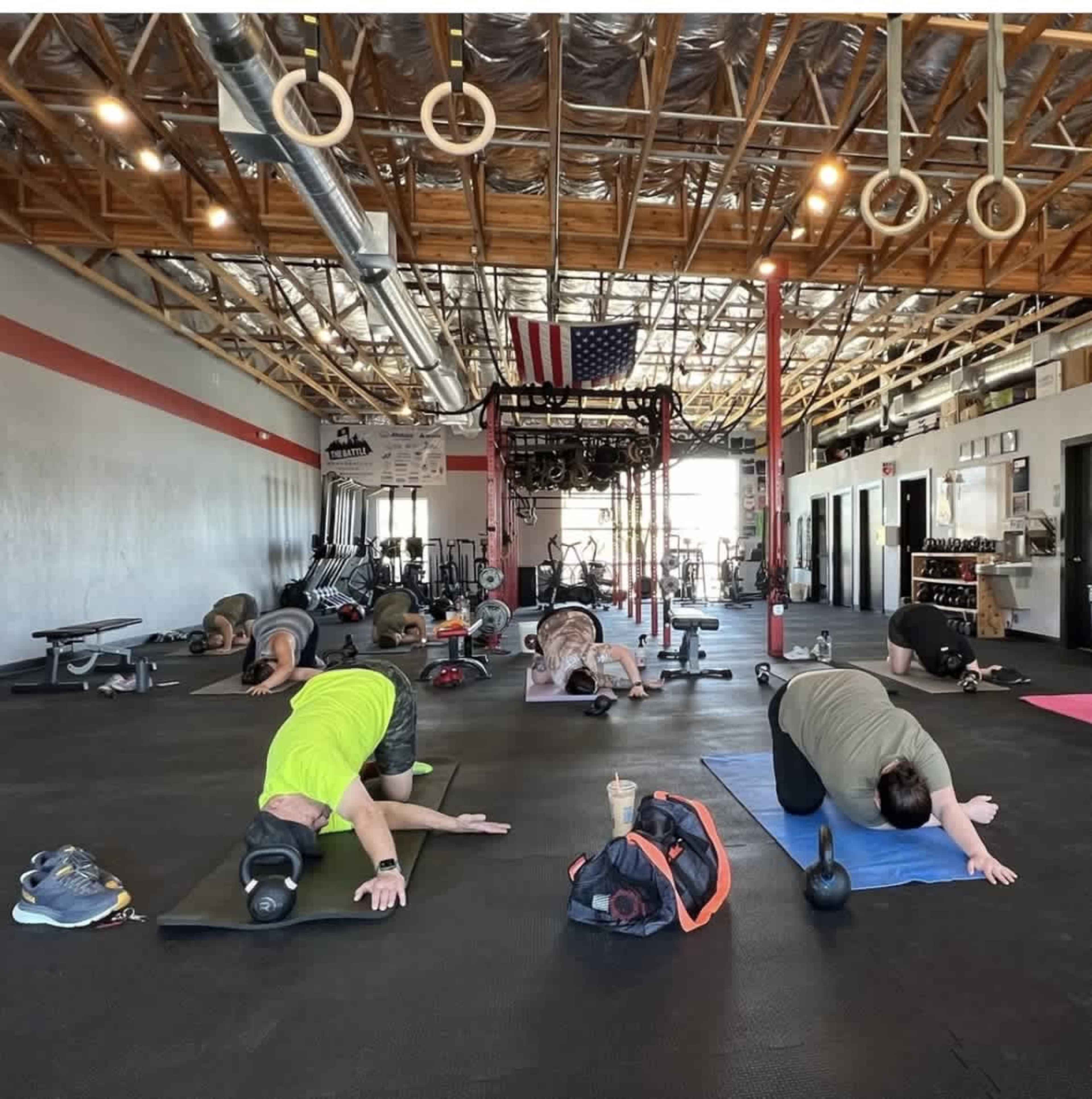 A group of people performs exercises on mats in a spacious gym with weights and fitness equipment visible in the background.