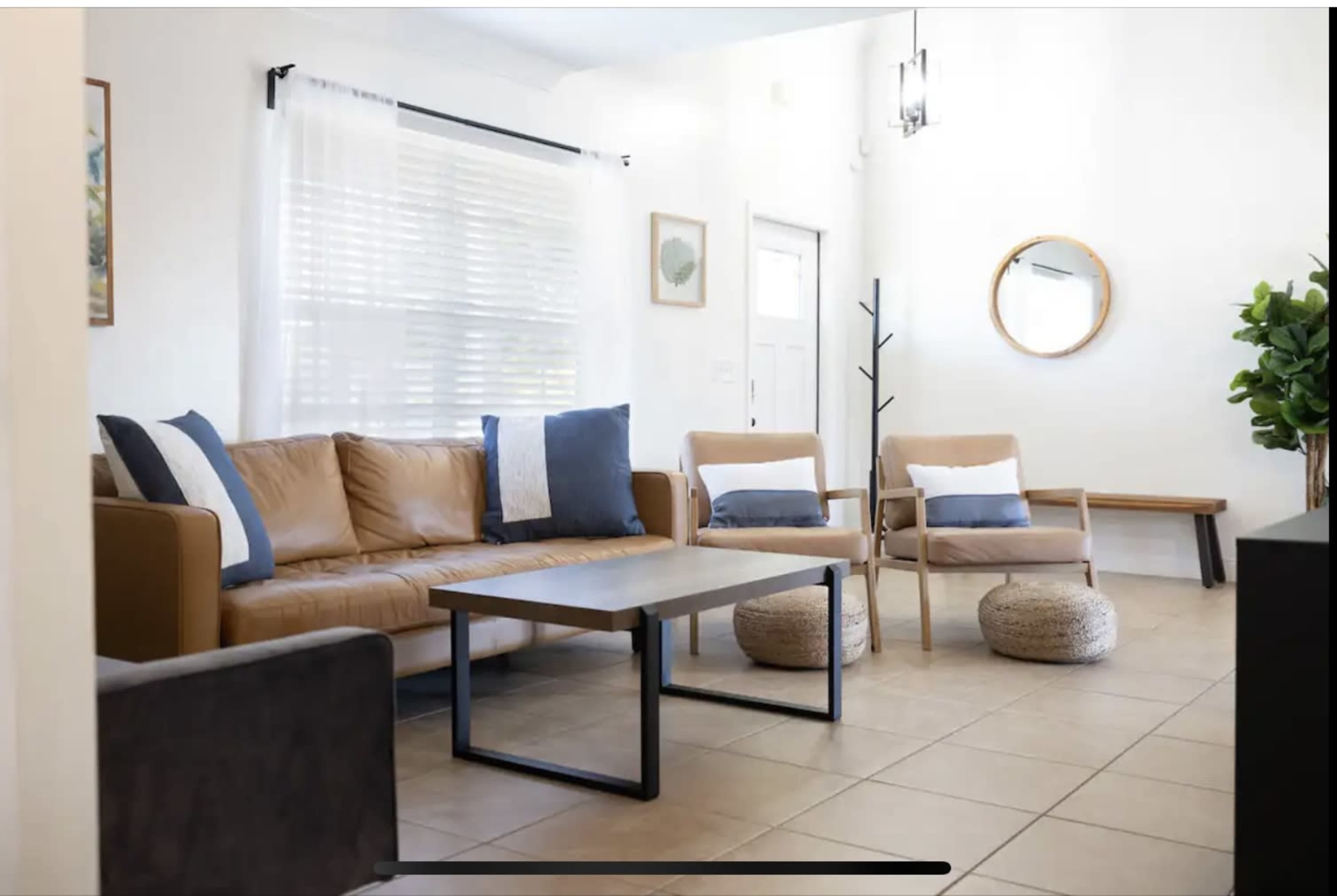 The image shows a modern living room with a tan leather sofa, two wooden chairs, a coffee table, and a round mirror on the wall.