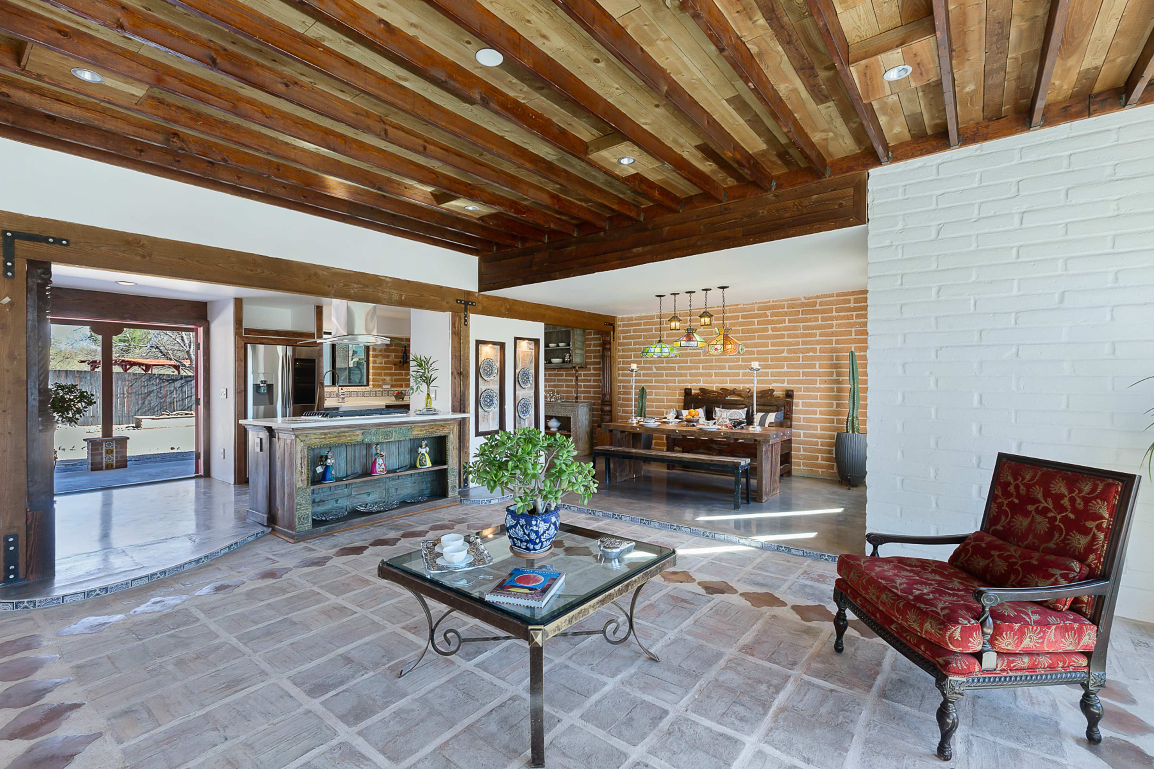 A spacious living area with wooden beams, a brick wall, and a tiled floor, featuring a coffee table and a red upholstered chair.