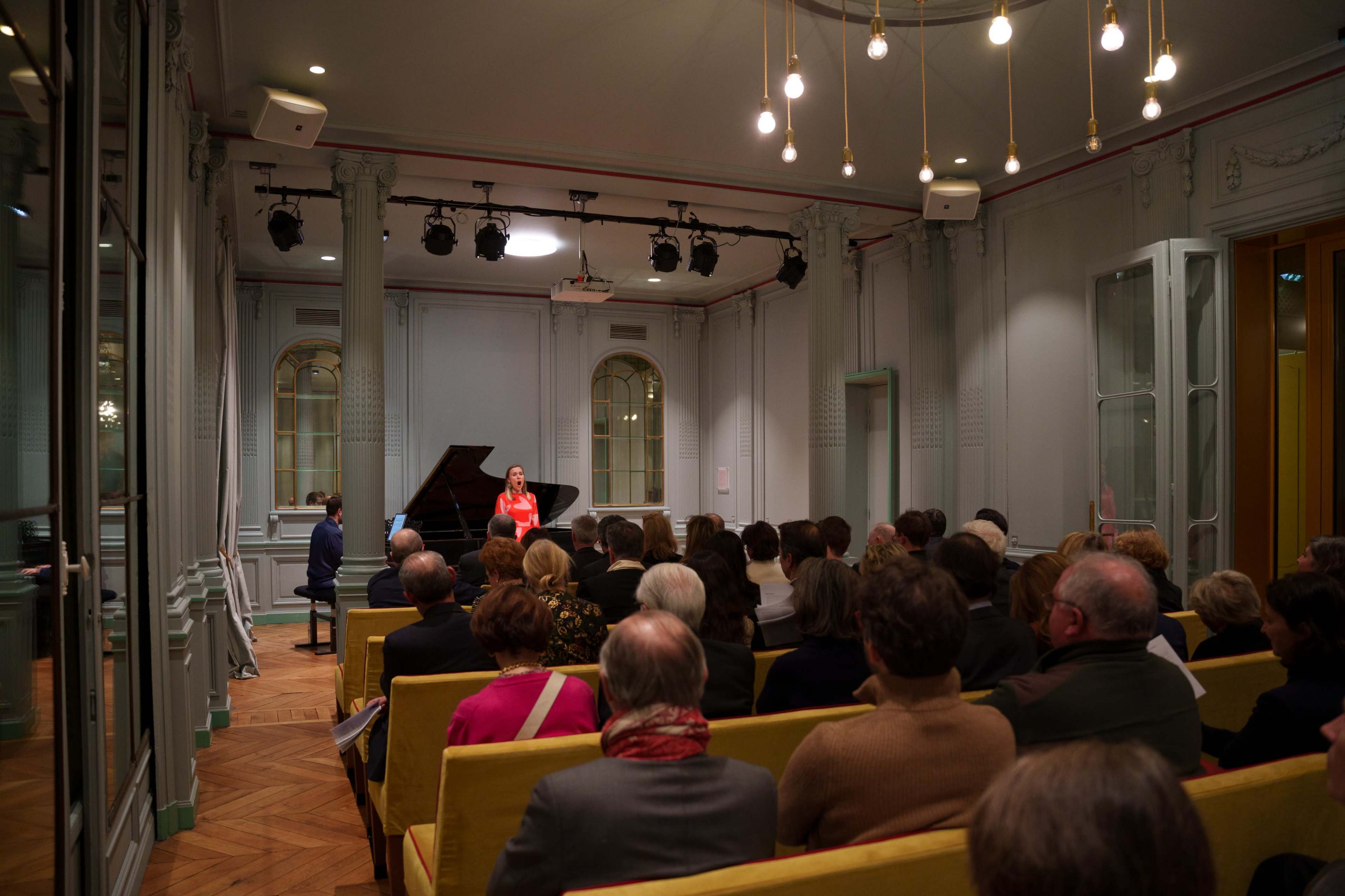 A pianist performs at a grand piano in front of an audience seated in a well-lit room with elegant decor.