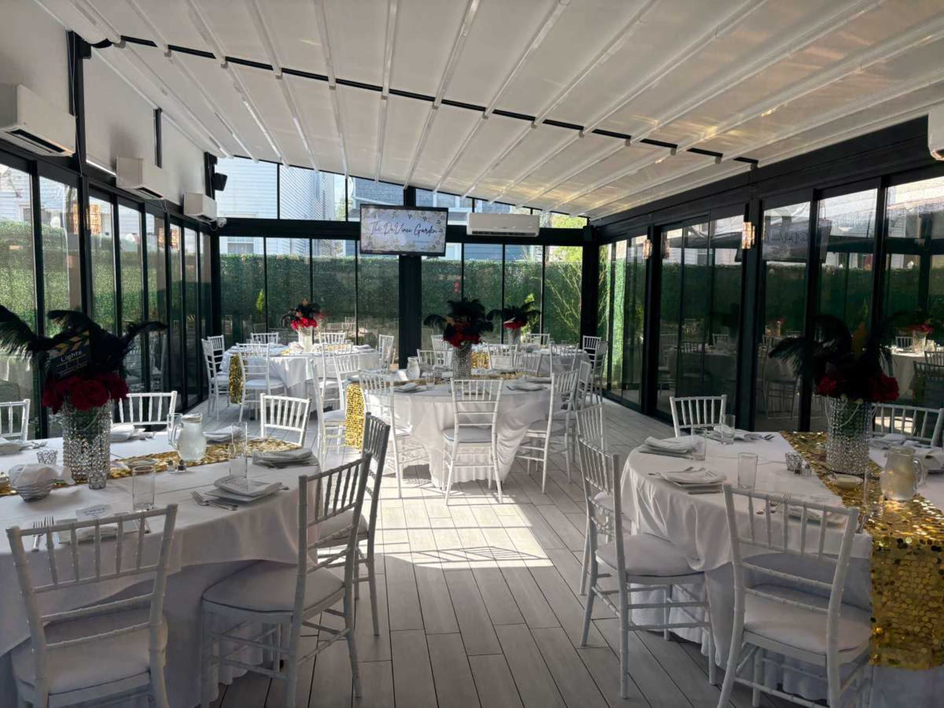 The image shows a brightly lit restaurant interior featuring white tablecloths, elegant chairs, and floral centerpieces, arranged for dining in a glass-enclosed space.