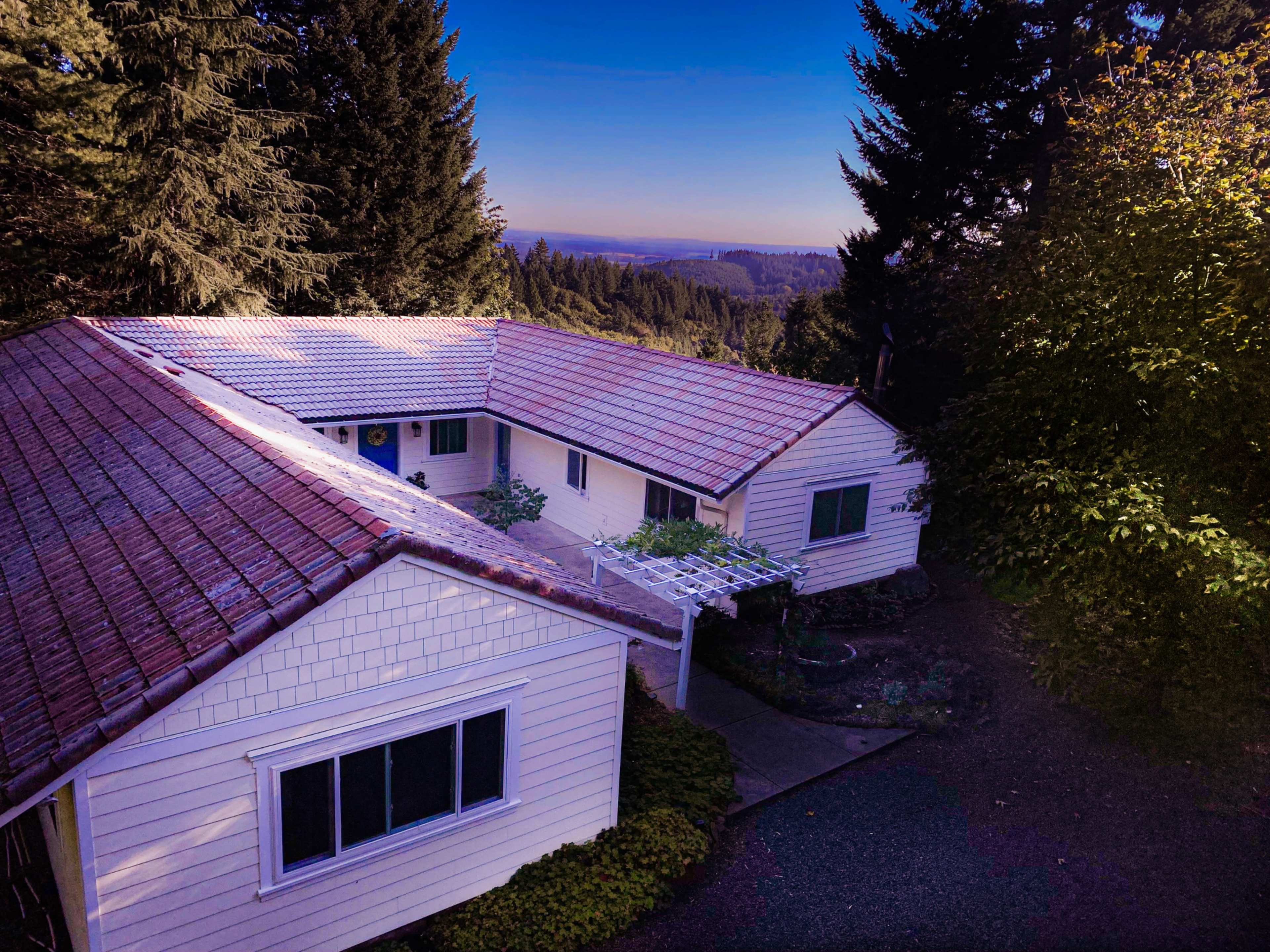 The image shows a single-story house with a tiled roof, surrounded by trees and a sloped landscape under a clear blue sky.