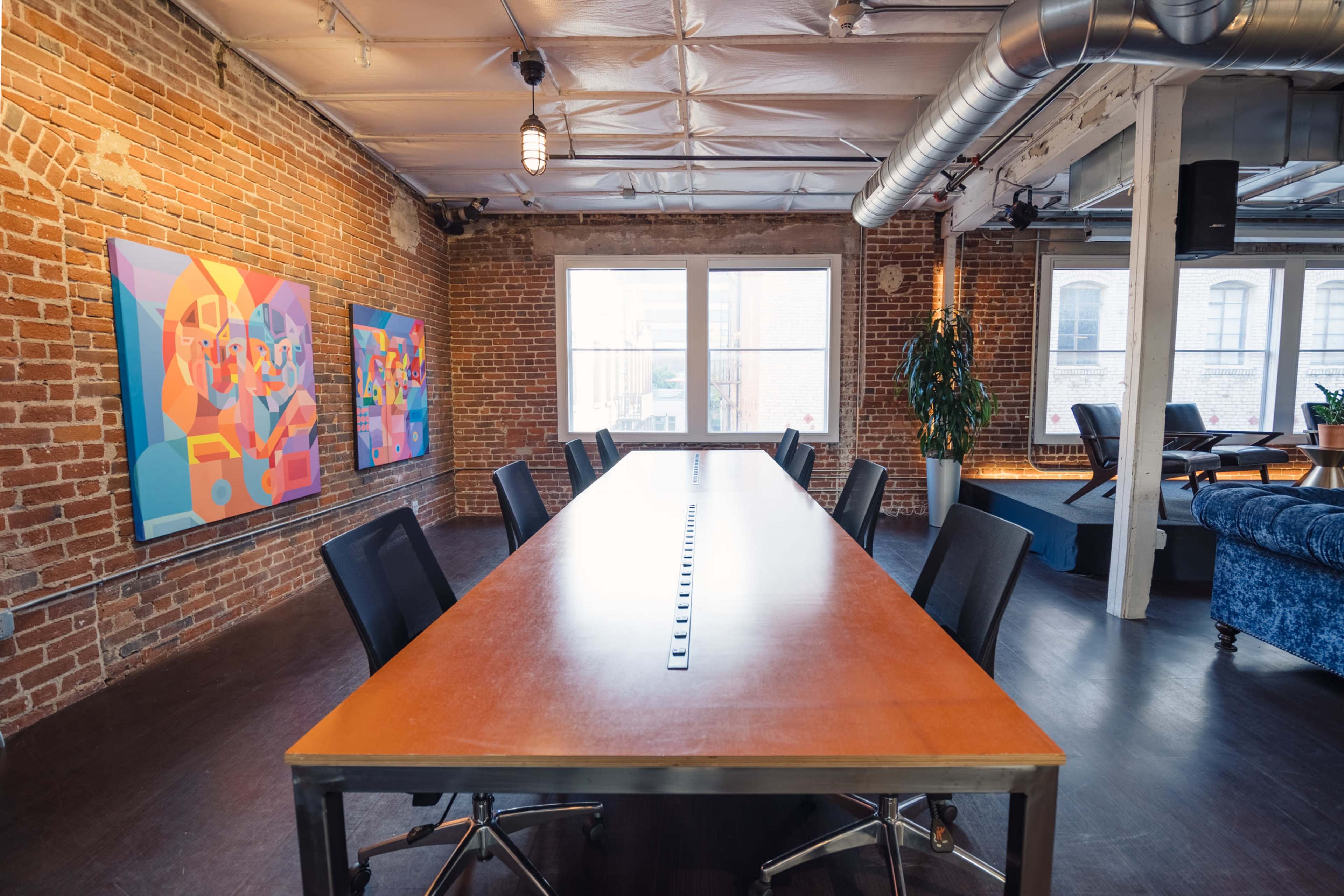 A large conference table is centered in a room with exposed brick walls and colorful artwork, featuring several windows that allow natural light to enter.