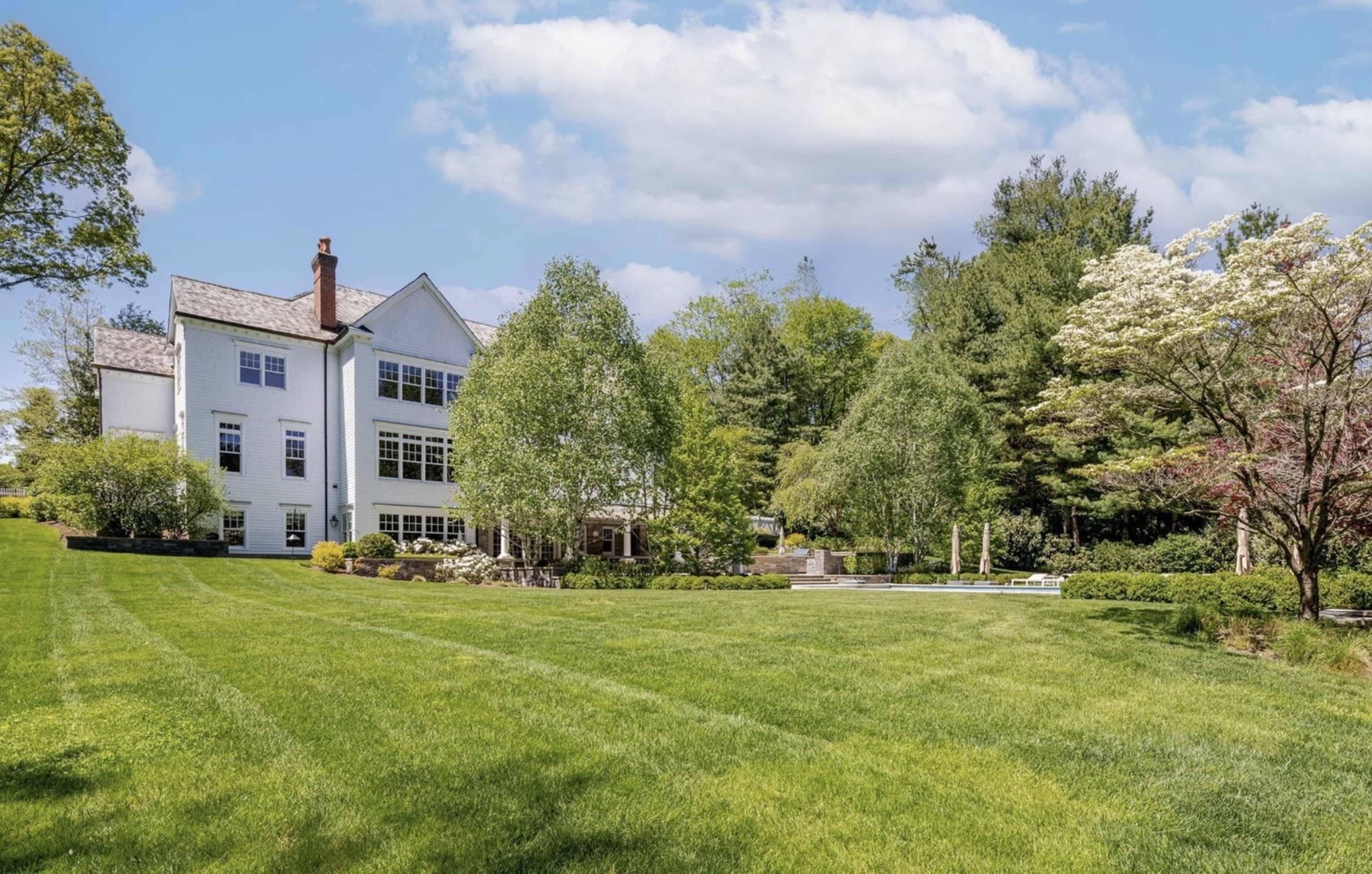 A large, white two-story house is set back from a lush green lawn, surrounded by trees and landscaped gardens.