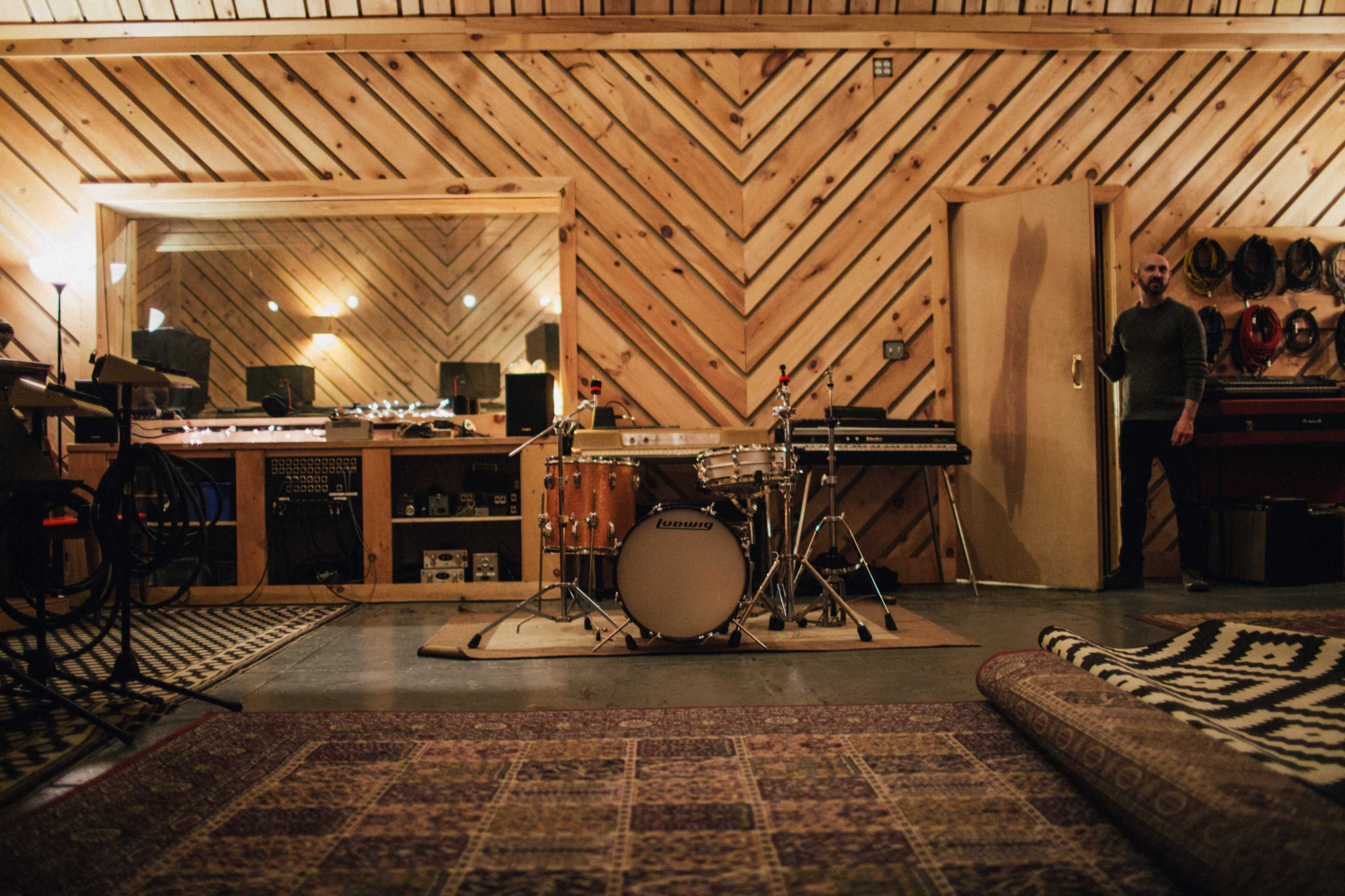 The image shows a music recording studio featuring a drum set in the center, wooden walls with diagonal paneling, and various electronic instruments and equipment in the background.